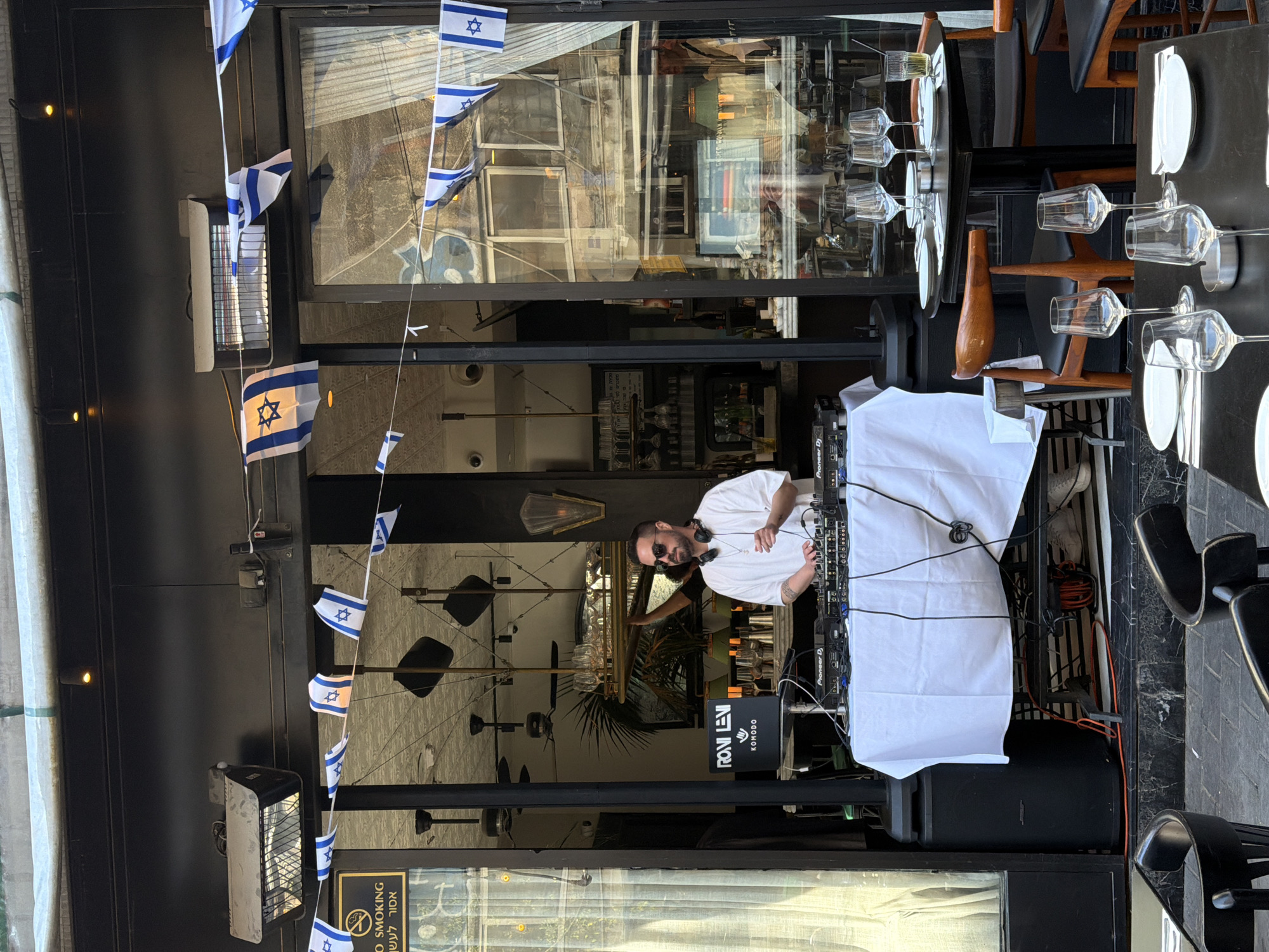 The image displays an outdoor or semi-outdoor cafe or restaurant scene, vibrantly decorated with strings of small Israeli flags. The setting suggests a lively atmosphere, possibly for an event or daily entertainment.
At the focal point, a male DJ stands behind a table draped in white, operating professional DJ equipment, including Pioneer DJ-branded mixers. He is dressed in a white t-shirt, wears sunglasses, and has headphones around his neck, appearing engaged with his setup. A large black speaker is positioned beneath the DJ table. Adjacent to the DJ setup, a black sign prominently displays "RONI LEVI" and "KOMODO."
The surrounding area is furnished with dining tables and chairs, ready for patrons. Some tables are black, while others feature wooden tops, set with wine glasses, water glasses, white plates, and cutlery. Overhead, outdoor heaters are mounted on the dark building facade. Large glass windows behind the DJ reflect portions of the urban environment, including buildings and the sky.
A sign visible on the left side of the frame includes the text "NO SMOKING" in English and "אסור לעשן" in Hebrew. The bright, clear lighting indicates it is daytime. The numerous Israeli flags and the Hebrew text firmly establish the location as Tel-Aviv, Israel, as confirmed by the provided context. The overall impression is that of an active and festive gathering at a cafe or restaurant.