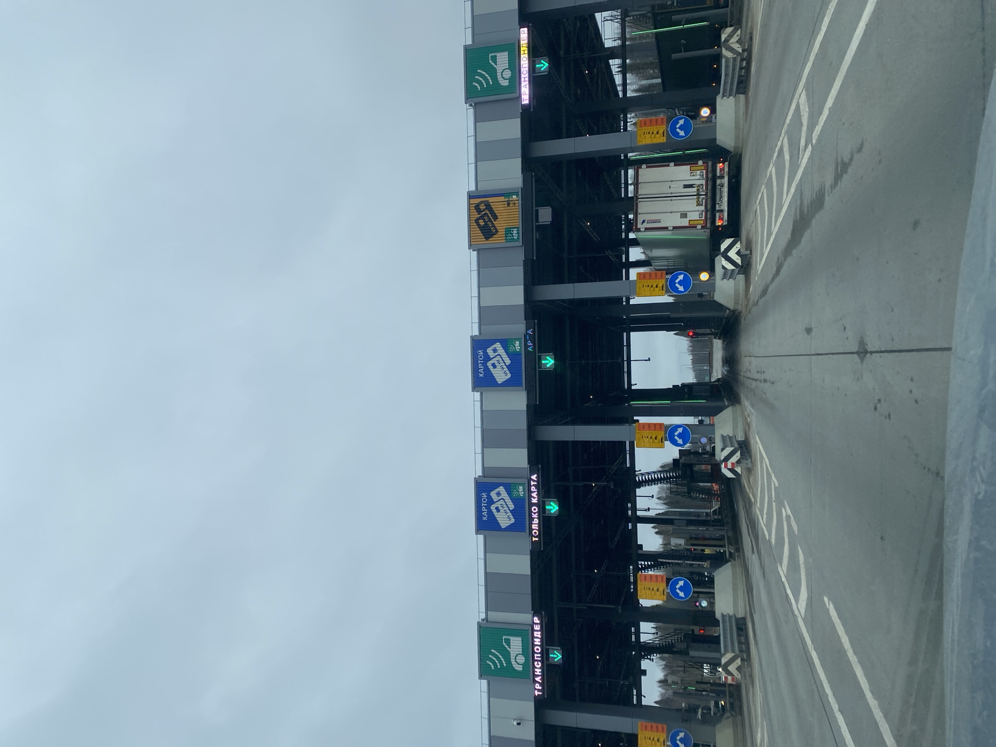 The image shows a toll plaza on a highway, likely located in Bizyar, Russia based on the Cyrillic text on the signs. The plaza is multi-laned with overhead signage indicating the payment methods accepted at each lane (transponder, card). The signage includes green arrows indicating open lanes. A truck is proceeding through one of the lanes. The sky is overcast, and the pavement is wet. The image appears to be taken from inside a vehicle approaching the toll plaza.