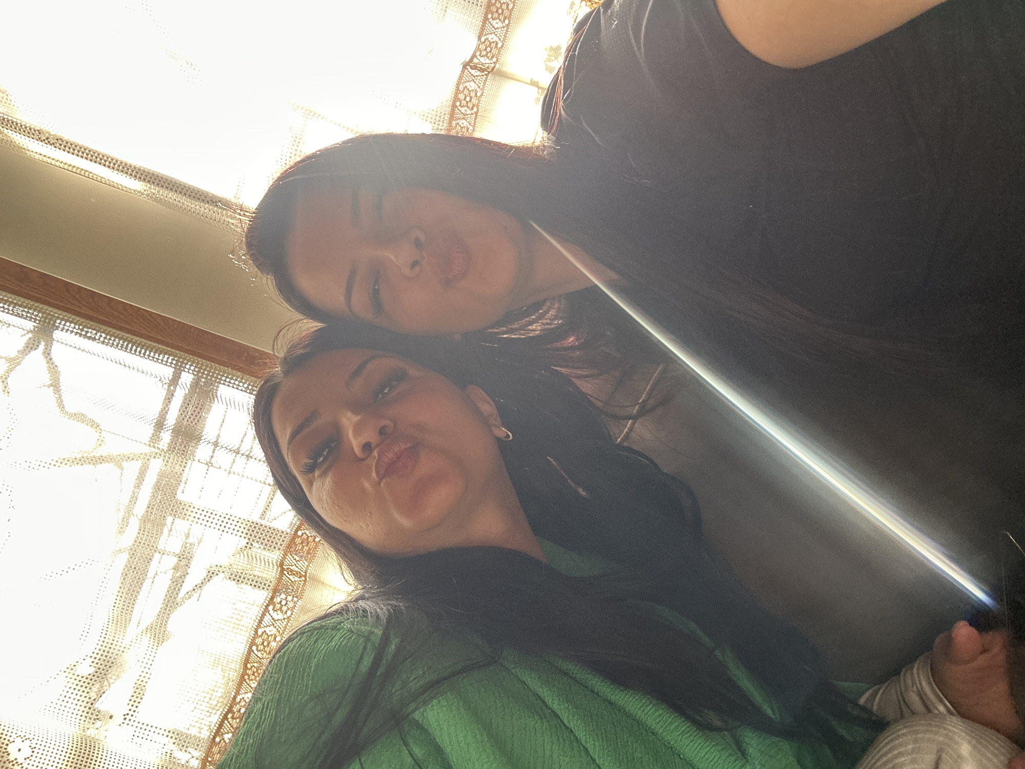 Two women are posing for a selfie. One woman is wearing a green top, the other is wearing a black top. They both have dark hair. The background shows a window with light coming through a lace curtain. The location is possibly Skala, Bulgaria.
