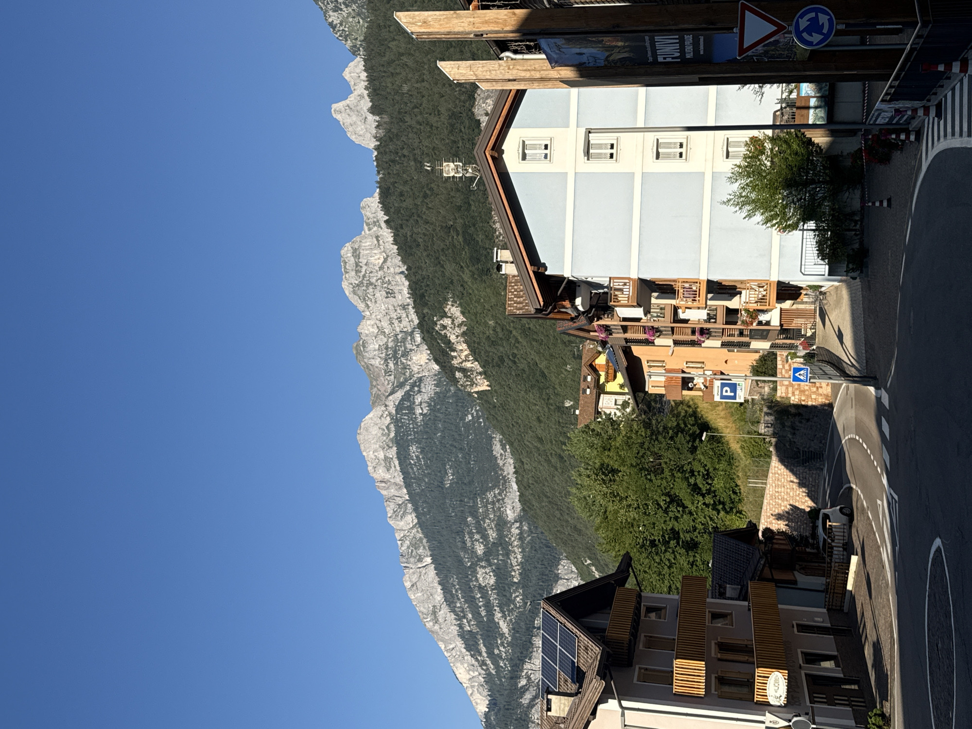 The image depicts a view of Molveno, Italy, on a clear day, showcasing a street scene with buildings and a mountain backdrop. In the foreground, a paved road curves to the right, featuring white lane markings and a pedestrian crossing. Several buildings line the street, varying in architectural style and color. To the left, a multi-story building with solar panels on its roof is visible, featuring wooden balconies with white railings. Adjacent to it, another building has a gray facade and a sign that is difficult to read.
A pale blue, multi-story building stands prominently on the right side of the street, with white-framed windows. To its right, a tall wooden post is present, with a sign reading "FUNIVIA" and some text below it. Traffic signs are also visible, including a yield sign and a roundabout sign.
Behind the buildings, a steep, tree-covered mountainside rises, with a rugged, rocky summit visible at the top. The sky is a clear, bright blue. A small white car is parked near one of the buildings. The scene appears to be a quiet, residential or commercial area within the town, with the impressive mountain range serving as a dramatic natural backdrop.