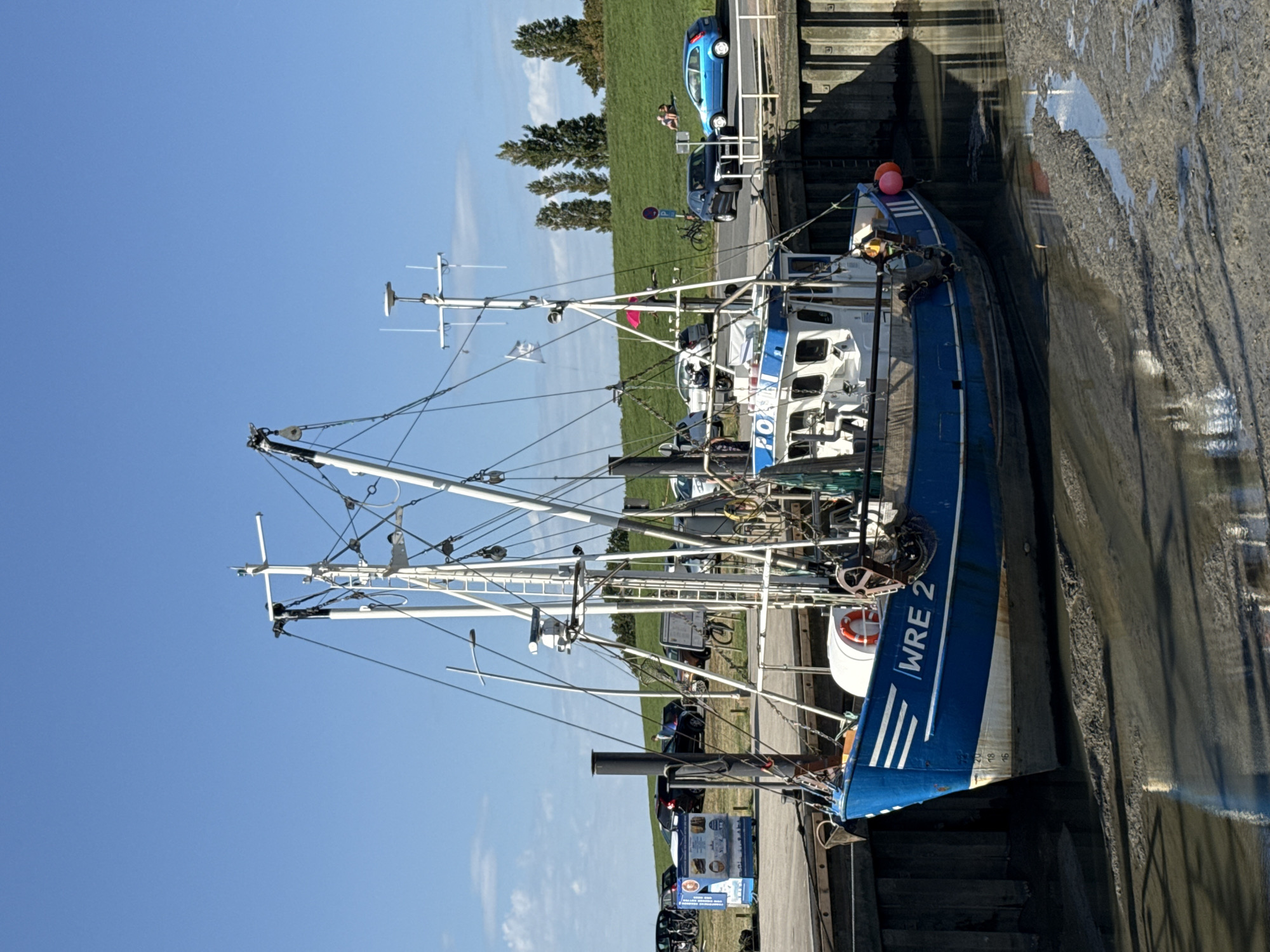 A blue and white fishing trawler, identified by "WRE 2" on its hull, is docked in a muddy harbor under a clear blue sky. The boat is equipped with a white mast, rigging, and various safety equipment including a life preserver and buoys. The foreground shows exposed mudflats, indicating low tide. In the background, a grassy embankment lines the harbor, with several cars parked and two individuals relaxing on the grass. A small blue car is visible on a road that runs along the embankment.