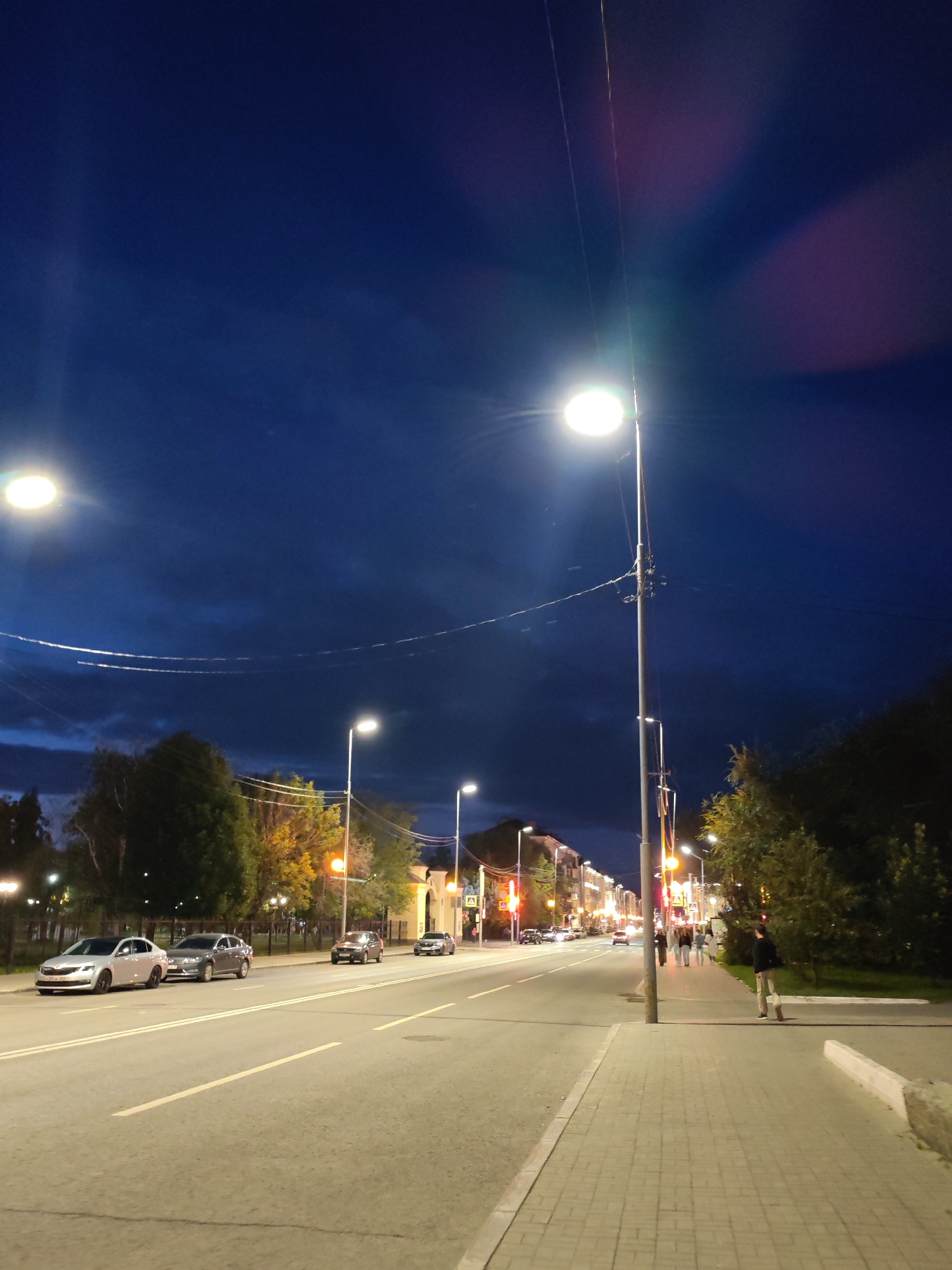 A street view captures a city scene at dusk or night. The sky is a deep blue with some dark clouds, and streetlights illuminate the road and sidewalk. Several cars are parked along the side of the road, and others are driving on the street. A few people are visible walking on the sidewalk. Trees line the side of the street, and buildings can be seen in the background. The lighting suggests it is nighttime.