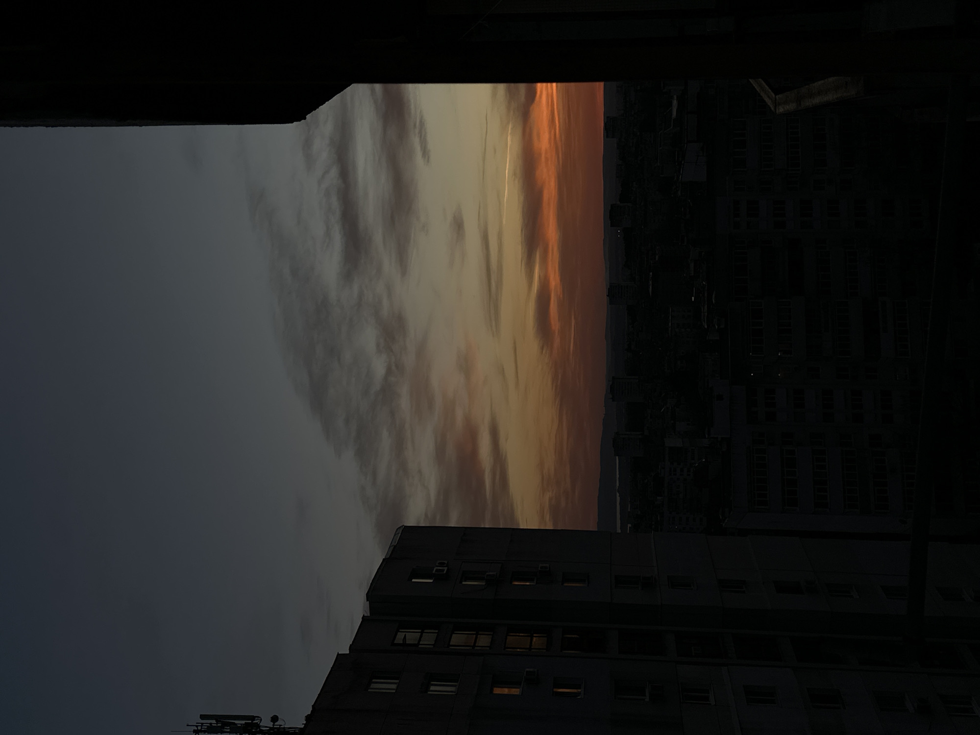 The image shows a view from a high vantage point, possibly a balcony or window, overlooking a cityscape during a sunrise or sunset. The sky is filled with clouds tinted with orange and yellow hues. Below the sky, dark silhouettes of buildings and the urban landscape can be seen. The framing suggests the view is partially obstructed by architectural elements. The building in front of the viewer has visible windows. The location is Varna, Bulgaria.