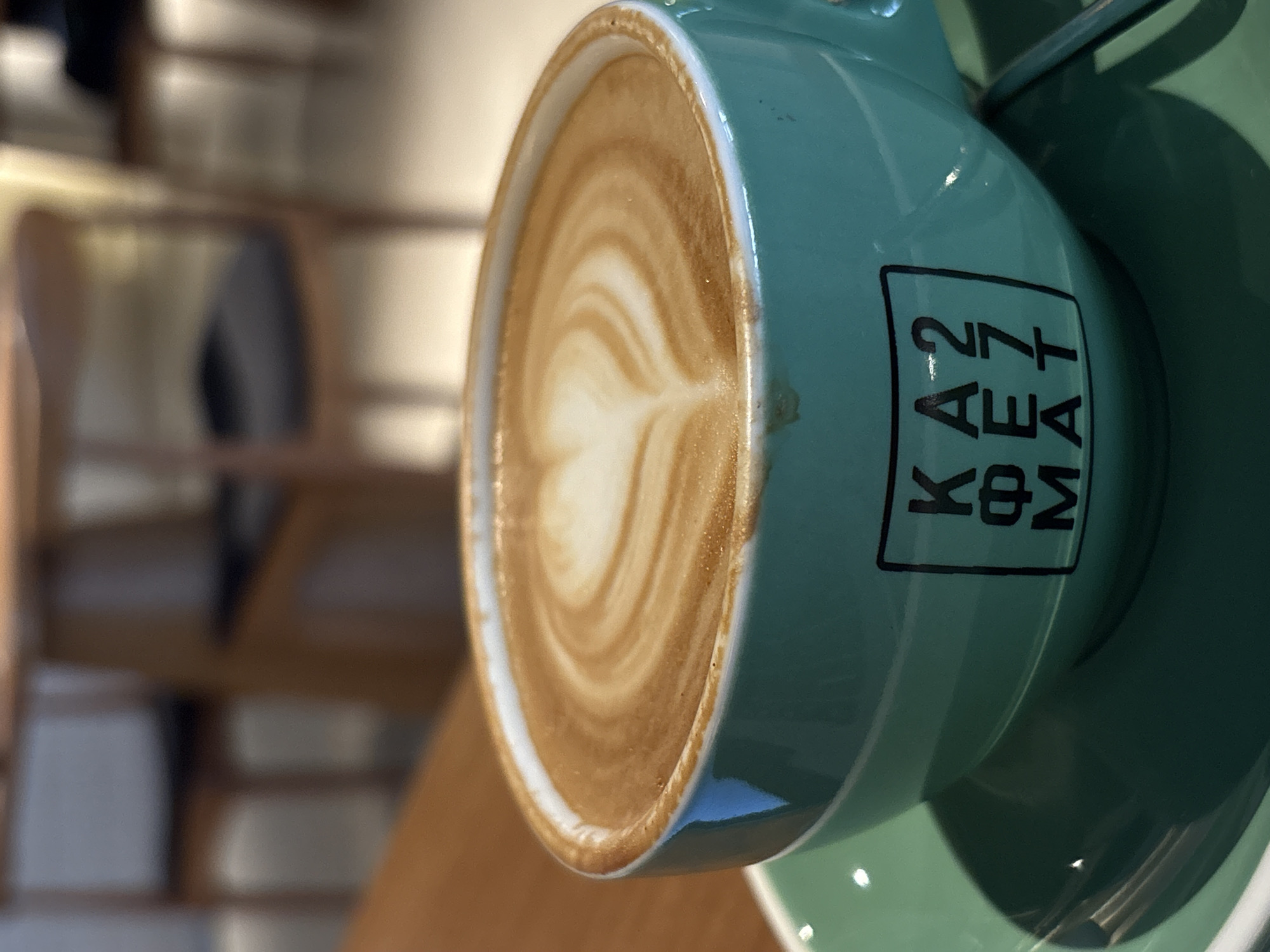A close-up, tilted shot depicts a light-blue ceramic coffee cup and saucer. The cup contains latte art in the shape of a heart. The cup has a logo written vertically on its side. The location appears to be a cafe in Kragujevac, Serbia, based on the logo "KAFEMAT" which is associated with a local coffee shop chain. The cup sits on a wooden table. The background shows a blurred view of chairs, suggesting an indoor cafe setting.