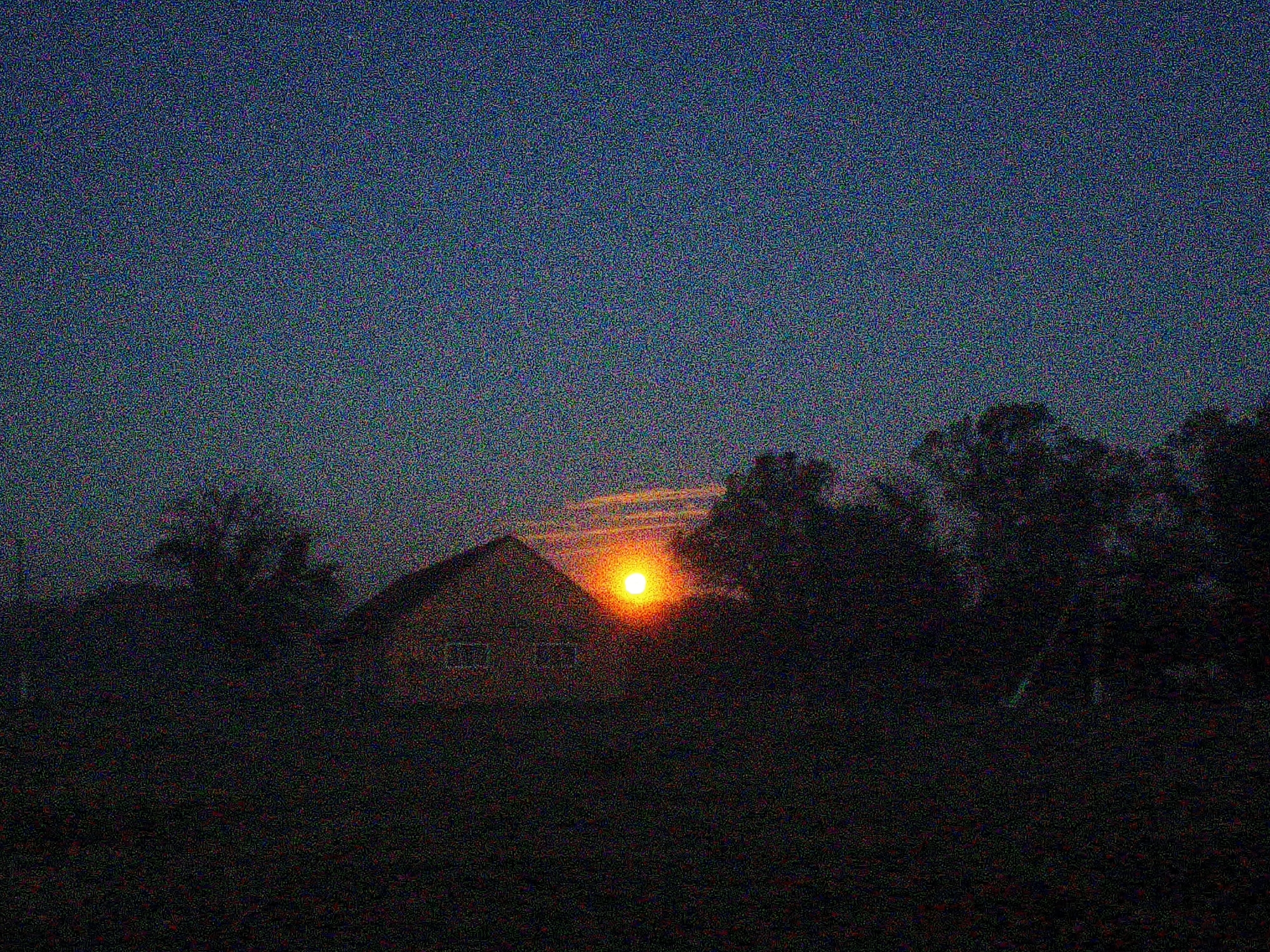 The image depicts a sunset scene. The sun is positioned on the horizon, obscured by trees and what appears to be a house or building. The sky is a gradient of dark blue to lighter hues near the horizon where the sun is setting. The foreground is dark, suggesting foliage or other objects in shadow. Due to the heavy noise and low resolution, the details are severely limited. There are no discernible people or actions visible. The environment is rural, judging by the foliage. There is no verifiable evidence to suggest that the location is specifically in Ustyvytsia, Ukraine, without more distinguishable clues or context within the image itself.