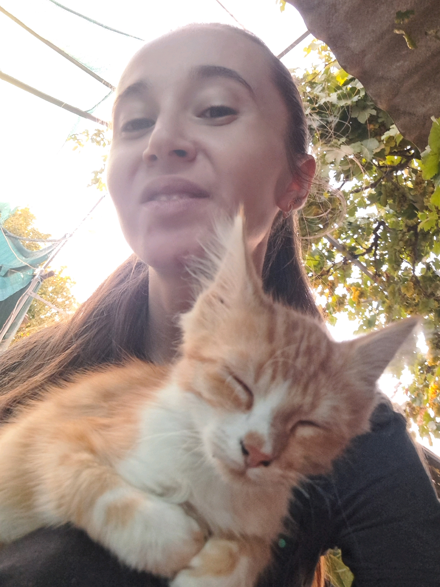 This is a selfie taken by a woman holding an orange and white kitten. The woman's face is in the upper portion of the frame, with the kitten resting on her chest and shoulder, taking up the majority of the lower half. The background suggests an outdoor setting with foliage and sunlight filtering through. The overall impression is a casual, personal moment captured in Vypasne, Ukraine.