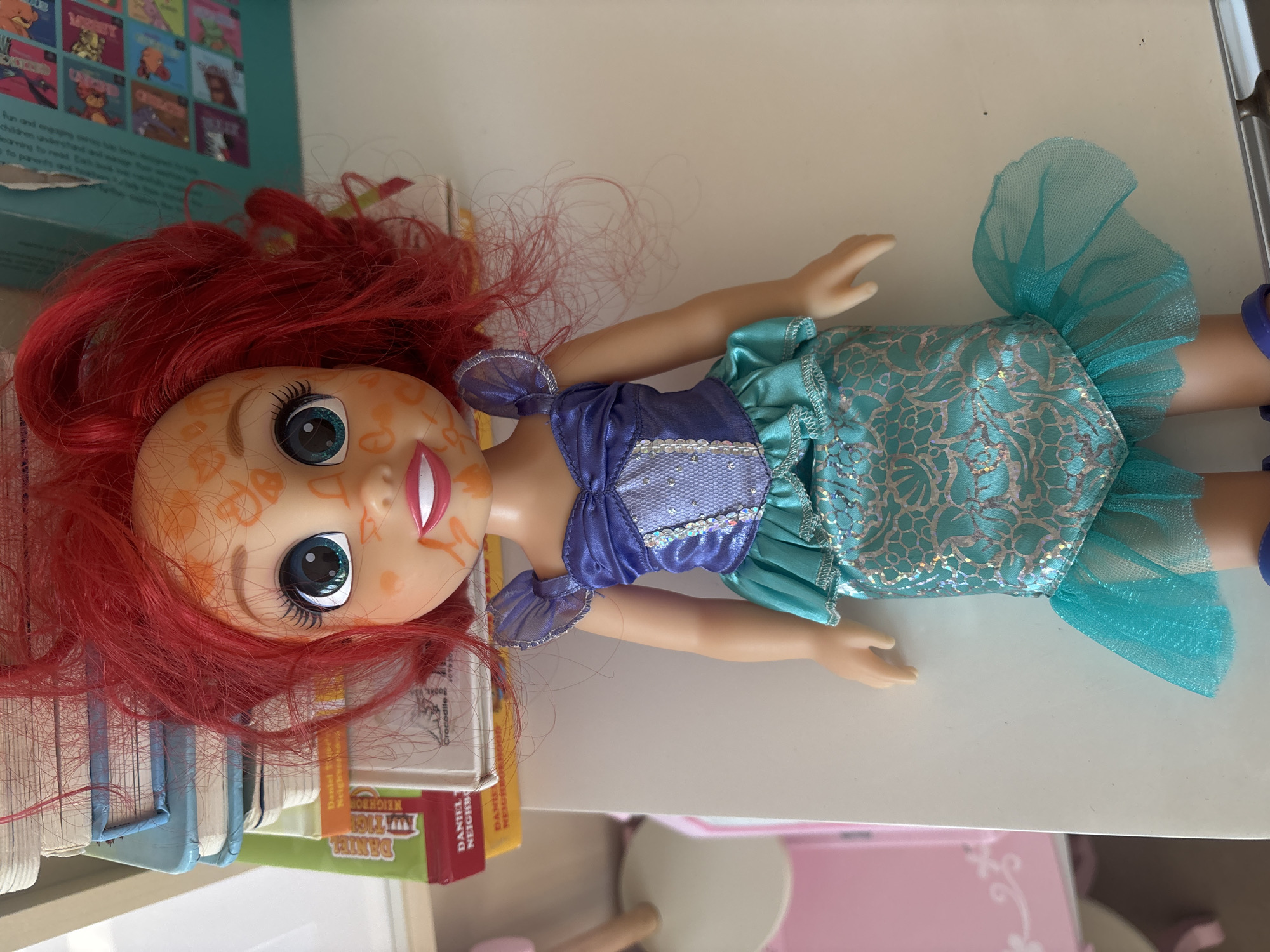 A doll with red hair, orange markings on its face, and a blue and turquoise mermaid outfit is lying on a white surface. Behind the doll is a stack of books. To the left of the doll is a pink table with a wooden leg. The scene is indoors.