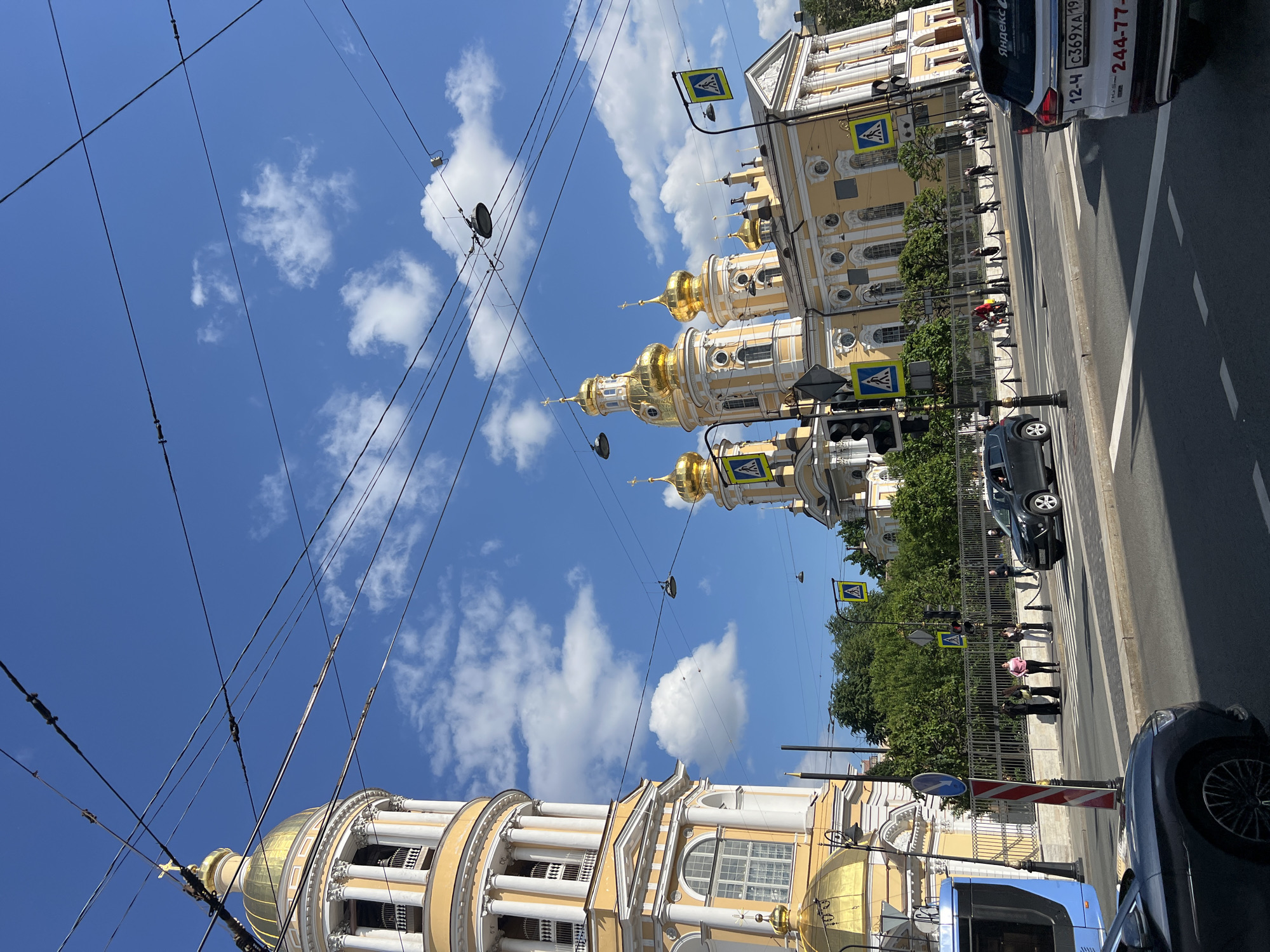 The image depicts a street view in Saint Petersburg, Russia, dominated by the yellow and gold domes of a prominent church, likely the Smolny Cathedral, given its architectural style and presence in the city. The church features multiple golden domes, ornate facades, and classical architectural elements such as columns and arched windows. To the left, a tall, cylindrical bell tower with a golden dome and white columns stands prominently.
The street in the foreground is a busy urban thoroughfare with multiple lanes of traffic. Several vehicles are visible, including a blue tram on the left, a dark SUV, and a white car with Russian license plates and markings suggesting it is a taxi or ride-sharing service. Pedestrians are present on the sidewalks and at a crosswalk, indicated by yellow pedestrian crossing signs.
Overhead, a complex network of electrical wires and streetlights crisscrosses the sky, a common feature in many cityscapes. The sky is bright blue with scattered white clouds, suggesting fair weather. Lush green trees line the background behind a black metal fence, separating the street from the church complex. The overall scene is one of a vibrant urban environment with significant religious architecture.