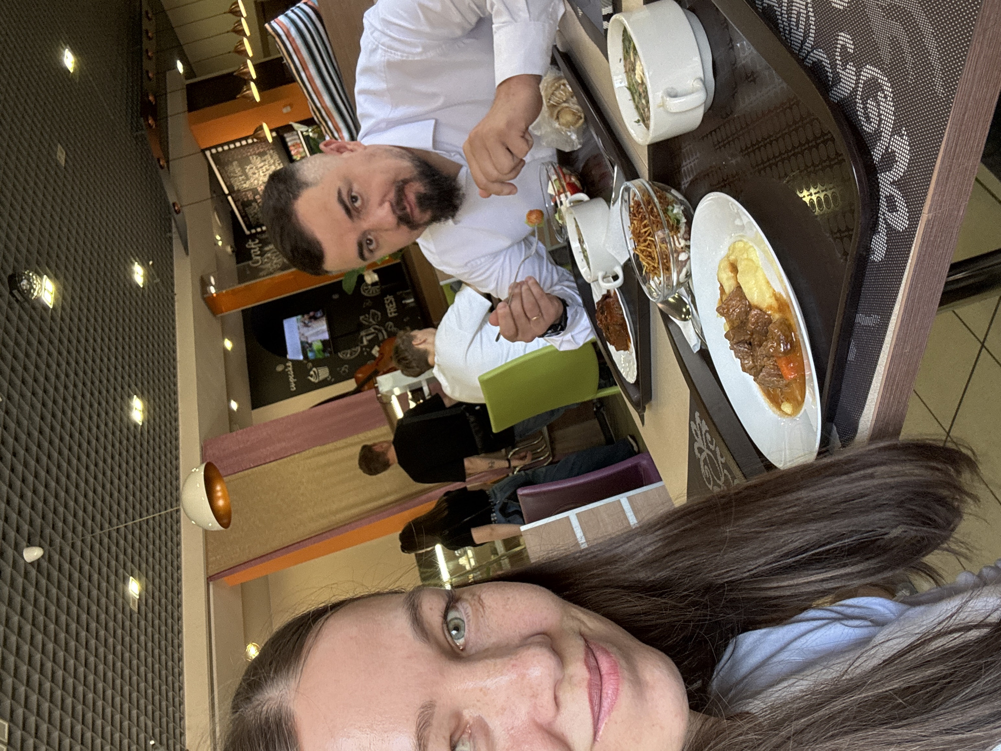 The image is a selfie of a woman with long brown hair, taken indoors, likely in a restaurant or cafe. A man with a beard and a white shirt is visible behind her, seated at a table. On the table are several dishes of food, including soup, salad, and a plate with meat and mashed potatoes. Other people are seated at tables in the background. The location is likely in Chaikovsky, Russia.