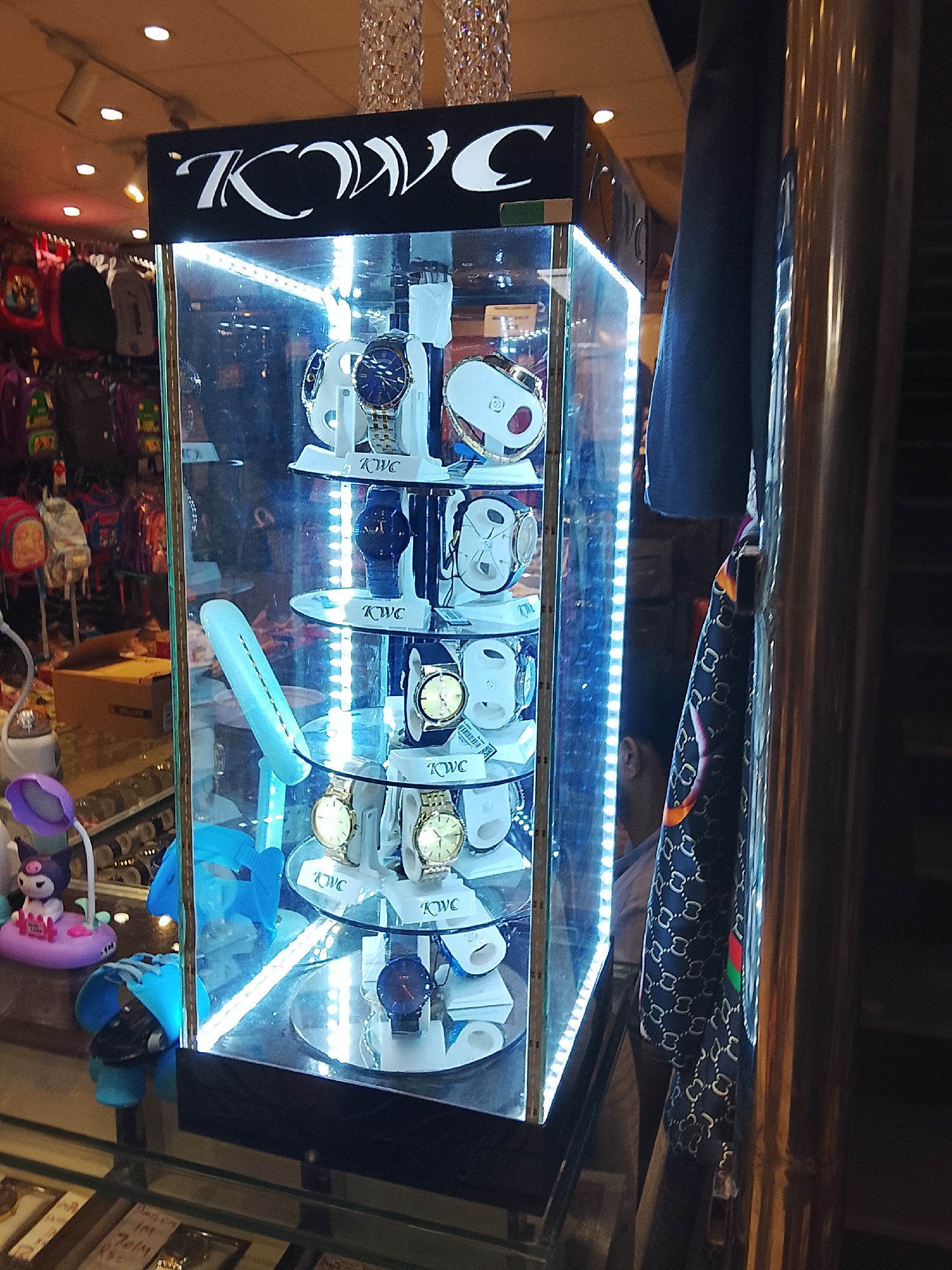 A glass display case filled with watches is in a store within Gulgasht Colony, Pakistan. The display case has the letters "KWC" at the top and contains five tiers of watches arranged on white stands. A blue lamp is on the left side of the display case. Other items, including what appears to be a toy, are on the shelf next to the display case. A bag with a repeated logo pattern hangs near the display case and a pole. Shelves with other merchandise, possibly bags, are visible in the background. A man is partially visible behind the display case.