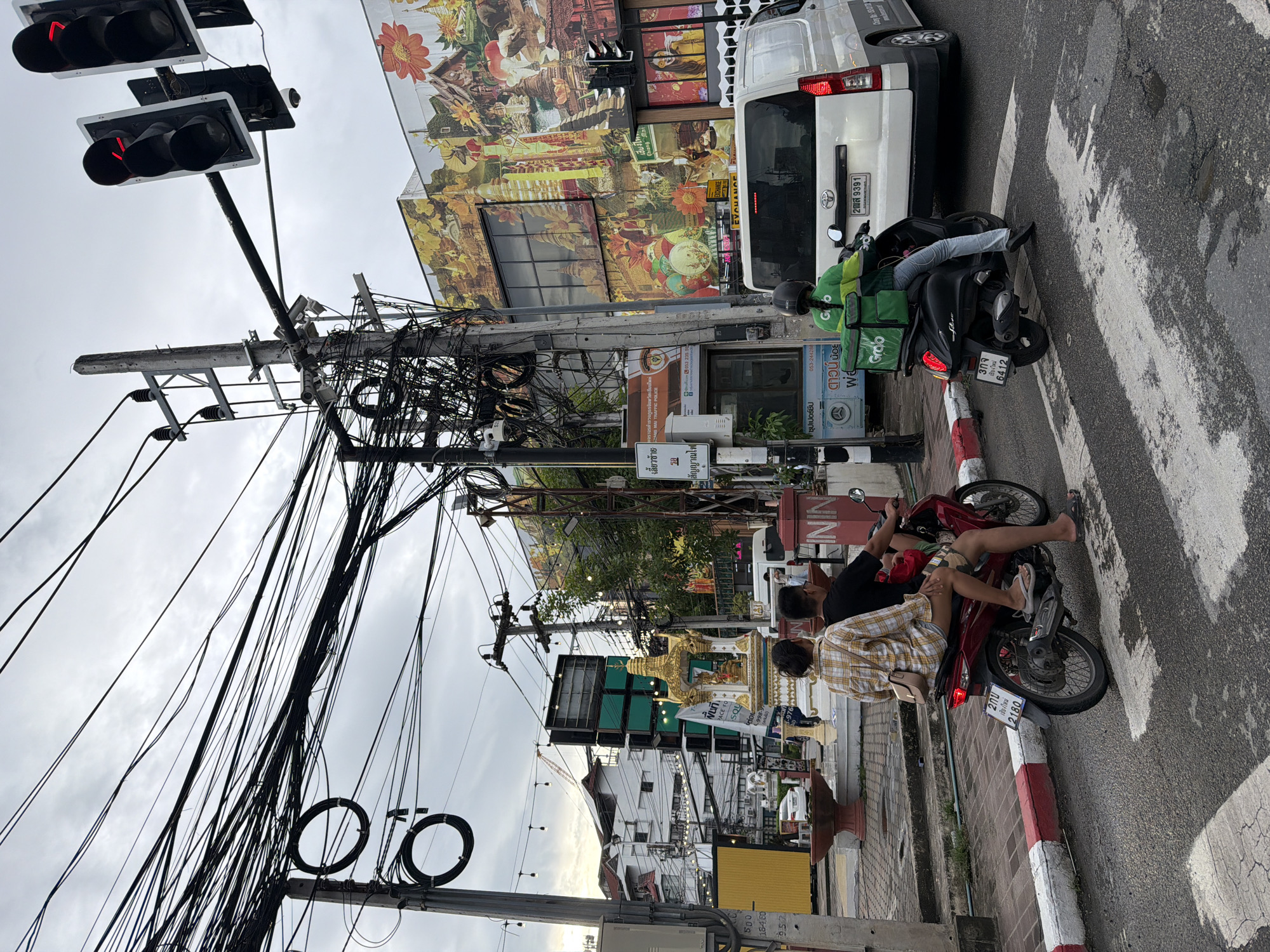 In Chiang Mai City Municipality, Thailand, on an overcast day, a busy intersection is depicted. Two people, a man and a woman, ride on a red scooter, crossing a pedestrian street. The woman wears a plaid shirt and the man wears a black t-shirt. Behind them, a person in a green jacket with a "Grab" logo on the back rides a black scooter. A white van is stopped at the intersection. The background features a building with a vibrant mural depicting temples, flowers, and scenes of Thai culture. Numerous electrical wires are strung overhead, a common sight in the city. Traffic lights are visible, with the red light illuminated. There is a small, ornate shrine on the side of the road, decorated with golden spires and offerings.
