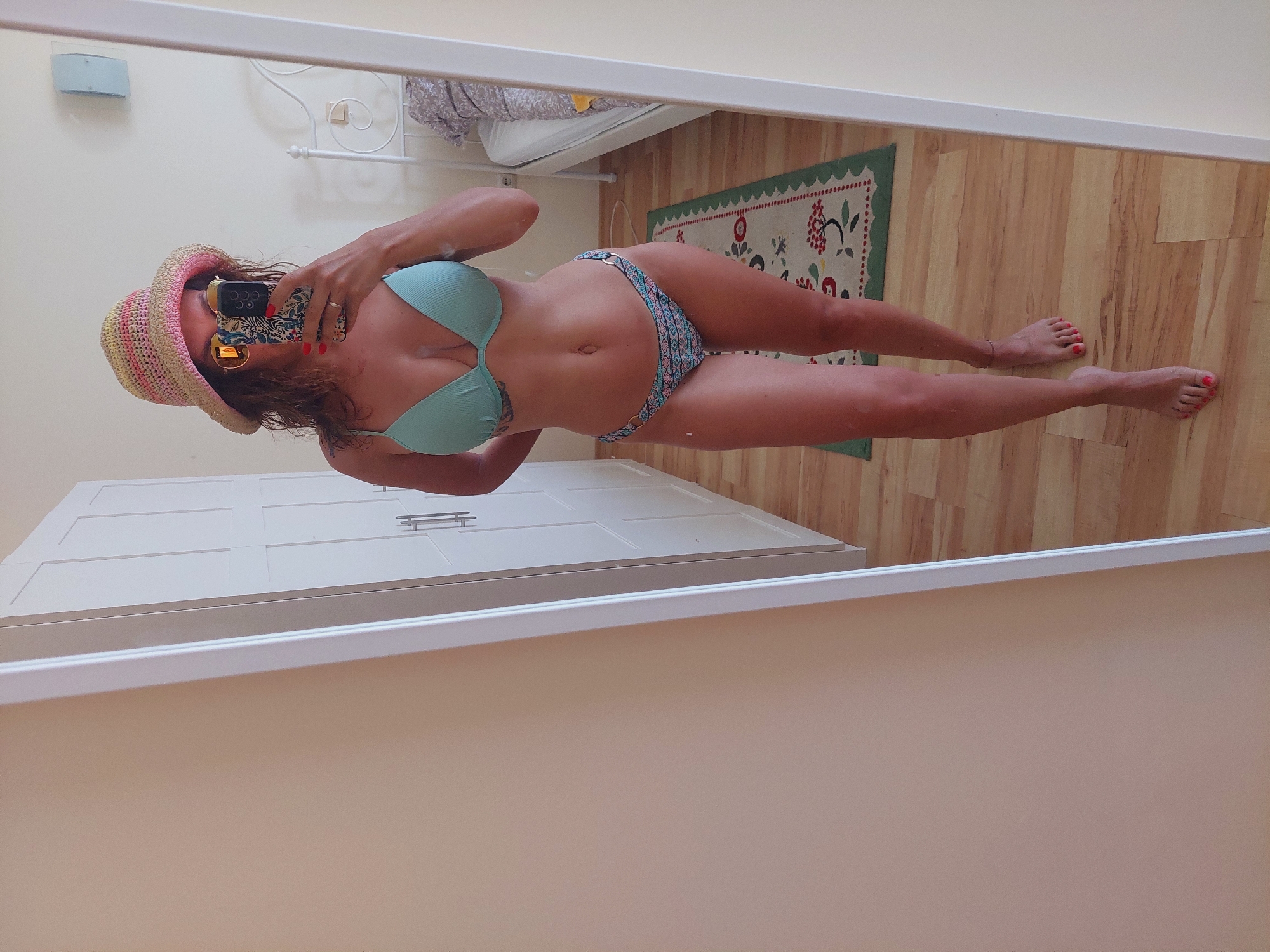 A woman wearing a light blue bikini and a straw hat is taking a mirror selfie in a room. The room appears to be a bedroom, with a white bed and a white wardrobe visible in the background. The floor is made of light-colored wood, and a small rug with a floral pattern is placed in front of the wardrobe. The woman is standing in front of a large mirror, which reflects her image. She is holding a smartphone in her right hand, and her left hand is resting on her hip. The overall lighting in the room is bright, suggesting it is daytime. The location is identified as being in the city of Sozopol, Bulgaria.