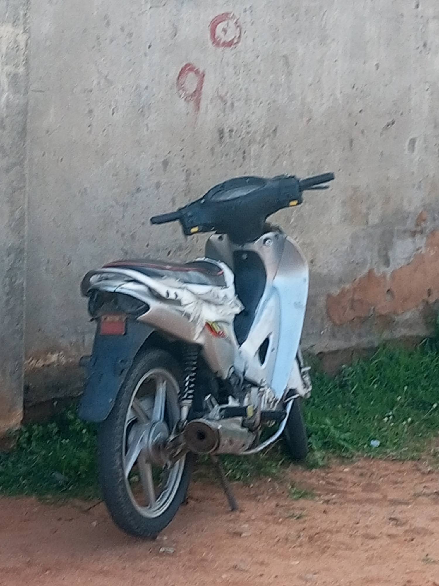 A silver and blue scooter is parked on a dirt path next to a rough, grey concrete wall with faded red markings. Green grass is visible to the right of the scooter, and the ground is covered in loose dirt and small pebbles. The scooter appears to be older, with some visible wear and tear on its body and seat. The overall scene suggests an outdoor setting in a somewhat neglected urban area, possibly in the city of Gombe, Nigeria. There are no people present in the image.