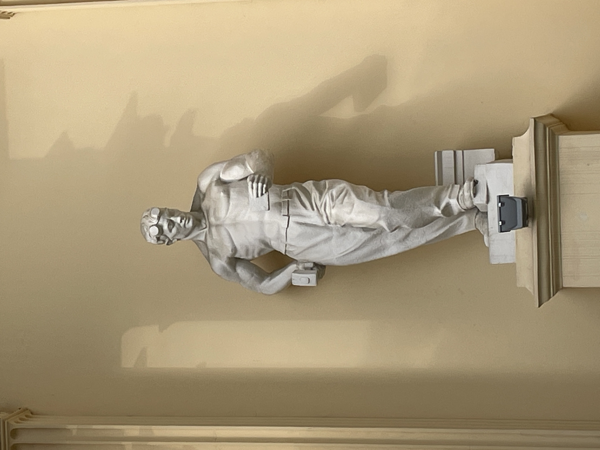 A statue depicting a male figure wearing goggles, possibly an aviator or pilot, is mounted on a wall bracket indoors. The wall is a light beige color and has molding along the bottom. The statue is positioned horizontally, lying on its side with its head facing left. This statue is on the ceiling above the main staircase, above the entrance to the Winter Theater in Sochi, Russia.