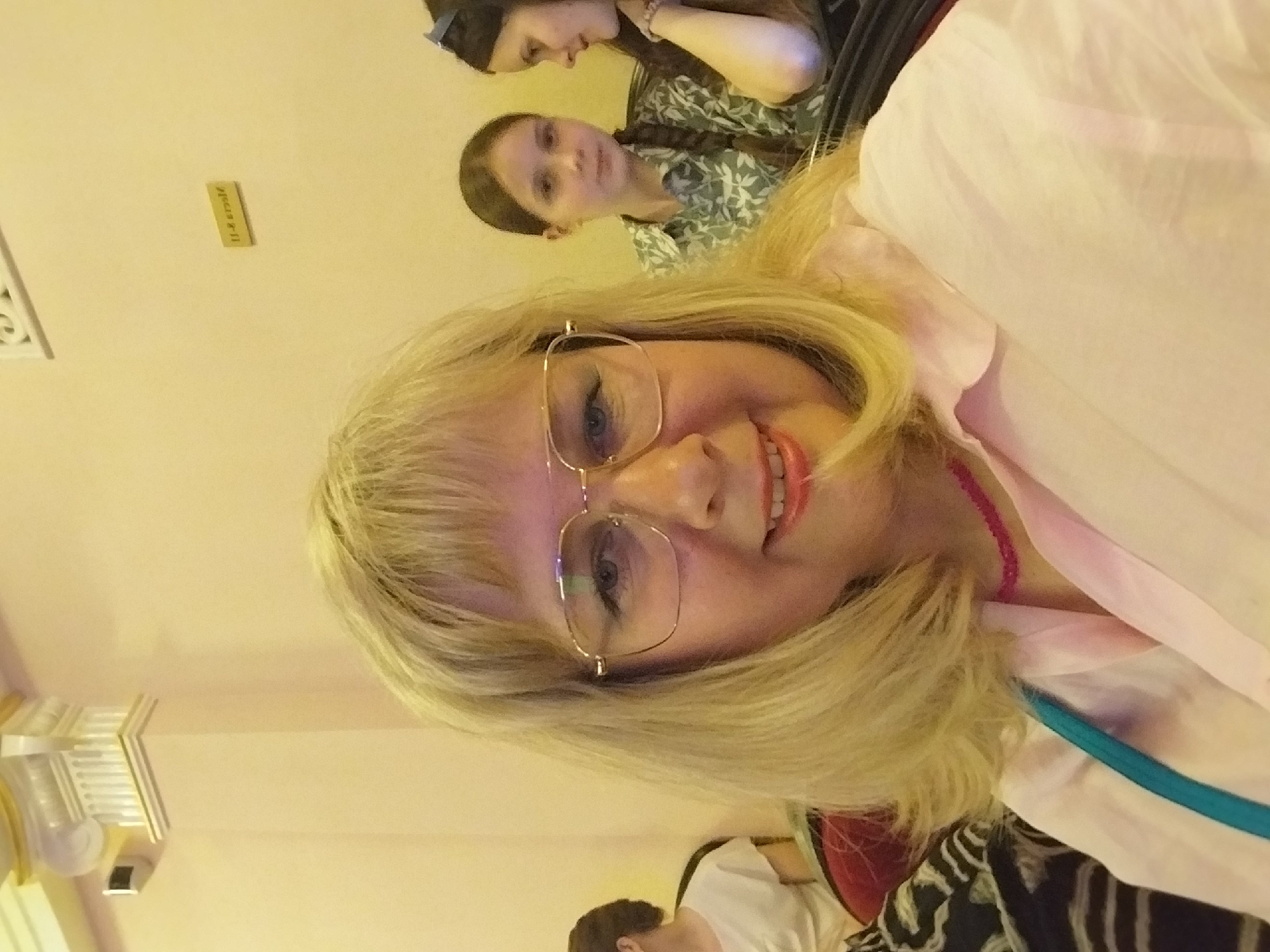 The image shows a selfie taken indoors. The main subject is a blonde woman with glasses and a pink necklace, looking towards the camera. Behind her, two other people are partially visible. One is a younger woman with dark hair in a braid, wearing a green floral shirt. Another person, partially obscured, has dark hair and is looking downwards. The setting appears to be a room with light-colored walls and some decorative elements visible in the background, including a metallic light fixture on the left. The overall lighting suggests it could be daytime or evening indoors. The mood seems casual and friendly, typical of a group selfie. A small rectangular object with yellow text is visible on the wall behind the people. The text appears to read "U.S. Embassy".