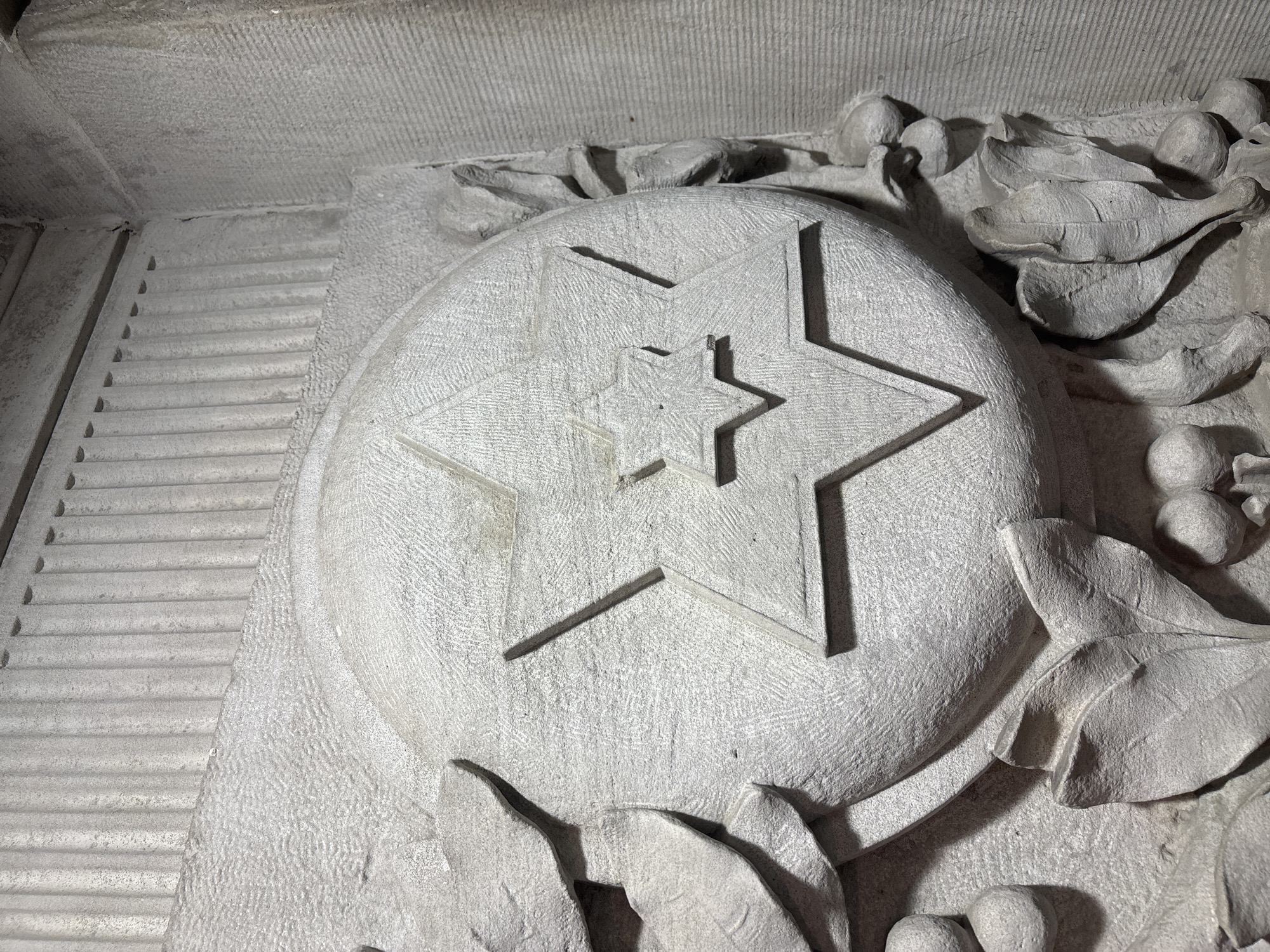 The image shows a stone carving of a five-pointed star within a circle, with a smaller star in the center. This carving is located on a building in Indianapolis, United States of America. The surrounding carving also includes stylized leaves and berries. To the left, there is a series of horizontal grooves. The stone appears to be a light gray color with a rough texture.