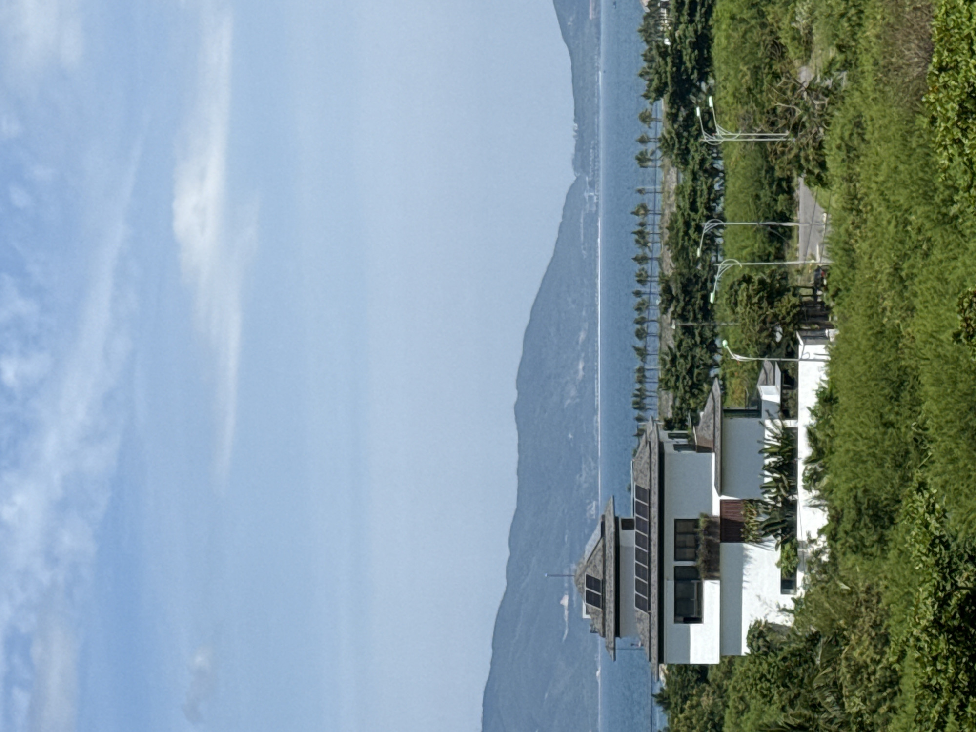 A bright, clear day is captured in this vertical, eye-level shot looking over a lush green landscape towards a body of water and distant mountains. In the foreground, a modern, white multi-story building with a dark, angled roof is partially visible, featuring several solar panels. The building is surrounded by dense green foliage, with tropical plants visible on its balconies and at its base. Beyond the building, a line of palm trees runs along the edge of a road, separating the greenery from a wide expanse of blue water. In the background, a range of blue-green mountains forms a scenic backdrop, disappearing into the hazy horizon. The sky above is a light blue, with scattered wispy white clouds. There are no people or animals visible in the scene. The scene appears to be a residential or resort area.