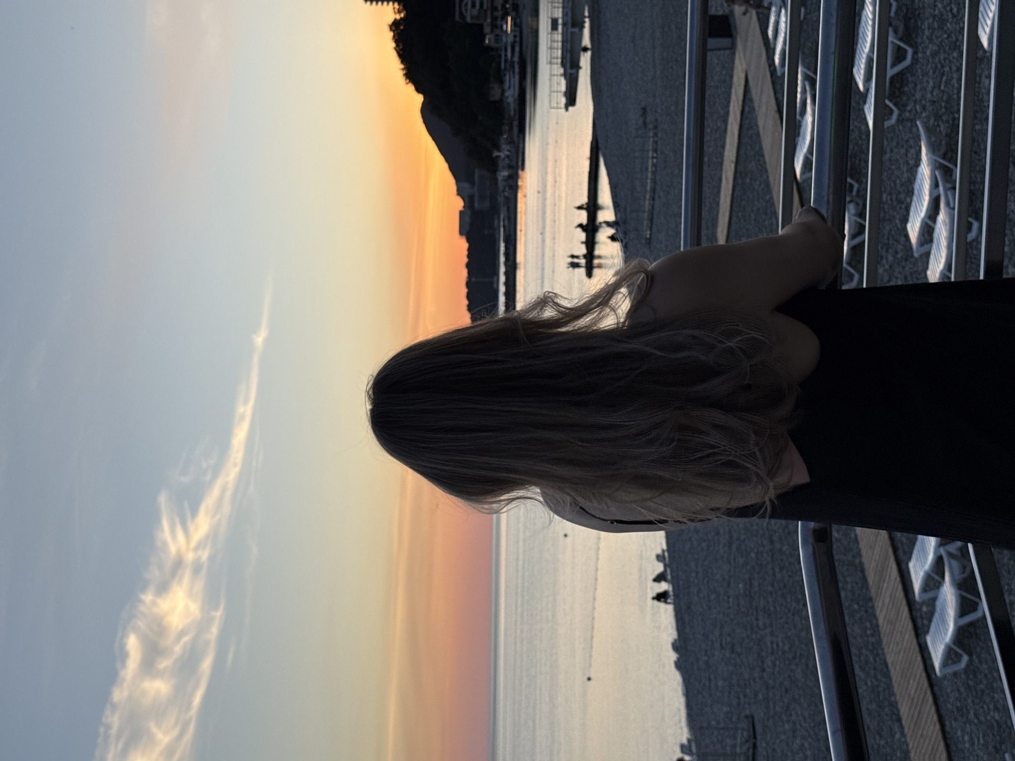The image captures a person viewed from behind, standing on an elevated platform and observing a sunset over a body of water in Nebug, Russia. The individual has long, light-colored hair and wears a dark, possibly sleeveless, garment, with their arms resting on a metallic railing. The sky displays a gradient of colors, from light blue in the upper portion to vibrant orange, yellow, and pink hues near the horizon, characteristic of a sunset, with wispy clouds present in the upper left. The water reflects these ambient sky colors. Below the railing, a pebble beach extends, featuring numerous white plastic sun loungers and sections of wooden boardwalk. Several indistinct human figures are visible as dark silhouettes on the beach and a pier in the midground. To the right, a dark, forested mountain slopes down to the coastline, revealing structures at its base and along its lower incline, and a small bay with boats and a dock is discernible in the mid-right.