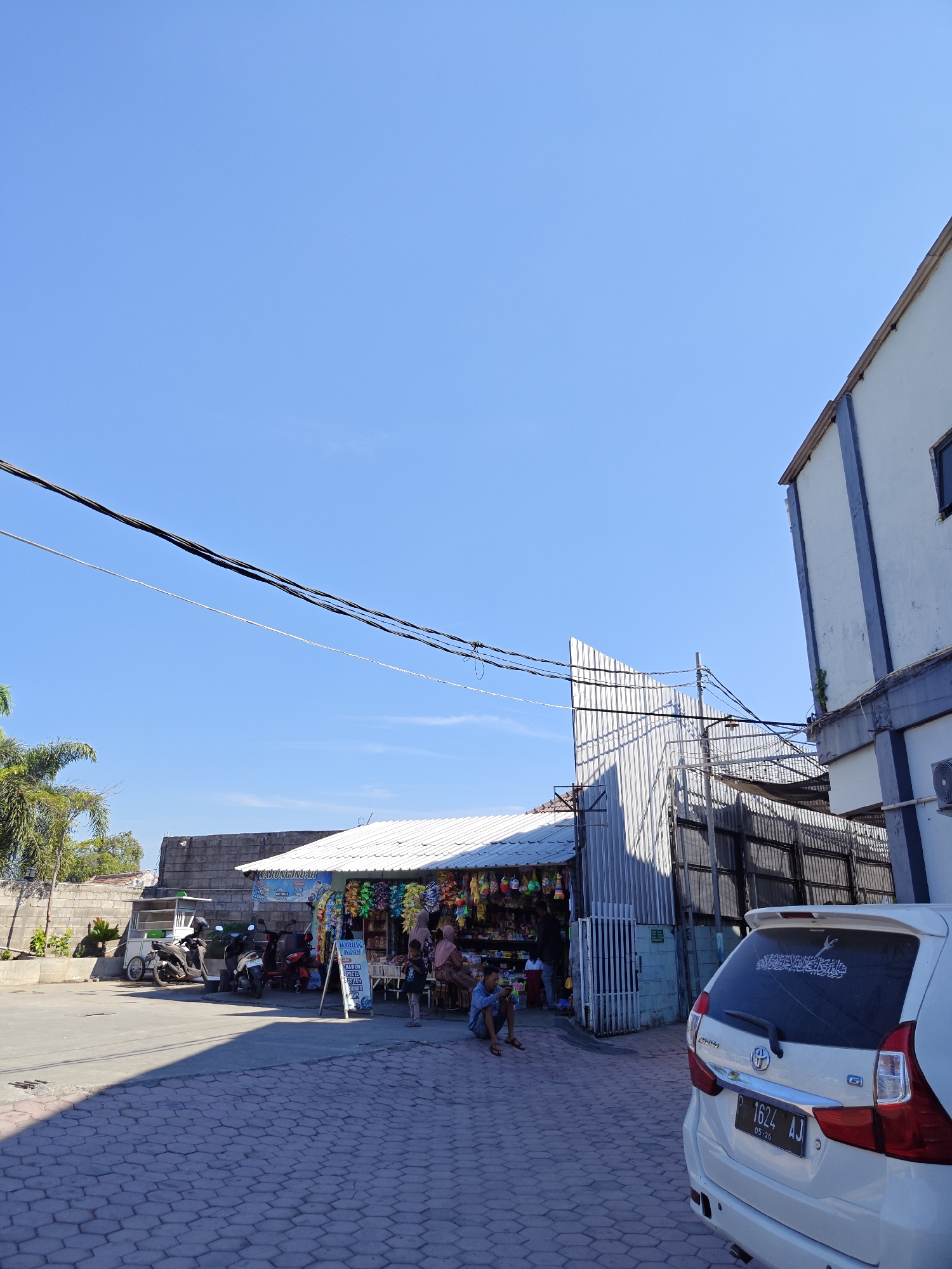 This image captures a street scene in Situbondo, Indonesia, under a clear blue sky. The foreground is dominated by a paved area with a hexagonal brick pattern. To the right, a white car is parked, its rear visible, with an Islamic calligraphy sticker on the back windshield and a license plate reading "P 1624 AJ".
In the mid-ground, a small shop with a white, sloped roof is set up. Various goods, appearing to be snacks or small merchandise, are displayed hanging from the front of the shop. Several people are present around the shop. Some are seated on the ground, including a person wearing a blue shirt on the left. Others are standing or interacting within the shop area. Several motorcycles are parked in front of a food cart and near the shop.
Behind the shop, a corrugated metal structure and a darker fence are visible. To the left of the shop, a low concrete wall and a brick wall form the boundary of the property, with a palm tree to the far left. In the background, a larger building with white walls and dark blue trim stands to the right, partially obscuring the sky. Overhead, power lines stretch across the frame. The overall impression is that of a typical, bustling local street in an Indonesian town.