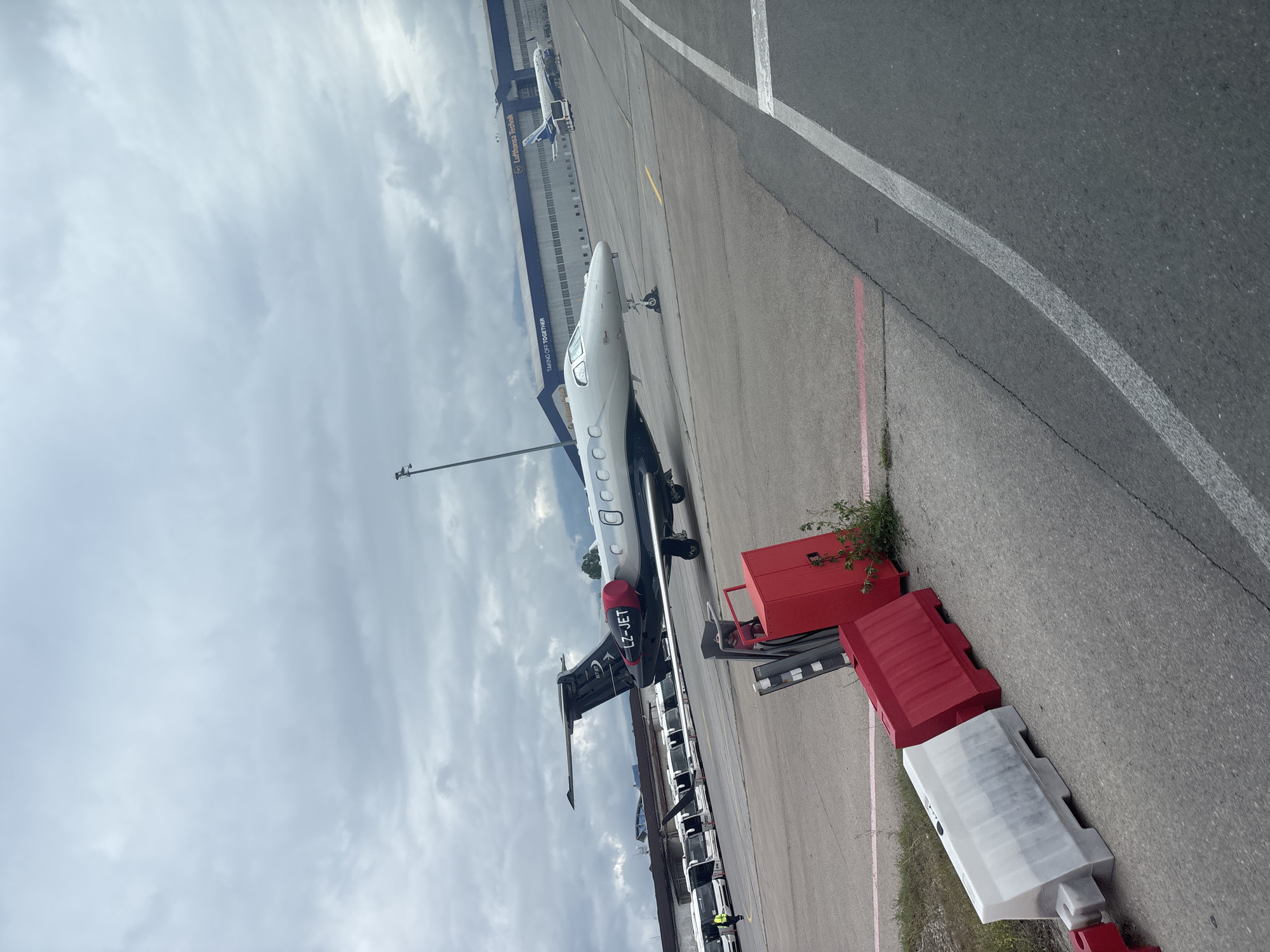 The image shows an airport scene. A white and black private jet is parked on the tarmac. The tail section of the plane features the registration "LZ-JET". Lufthansa Technik is visible in the background, indicating the location is likely Sofia Airport in Bulgaria. There are several small white airport service vehicles parked along the edge of the tarmac near the plane. The sky is overcast. Red and white safety barriers are positioned near the edge of the tarmac.