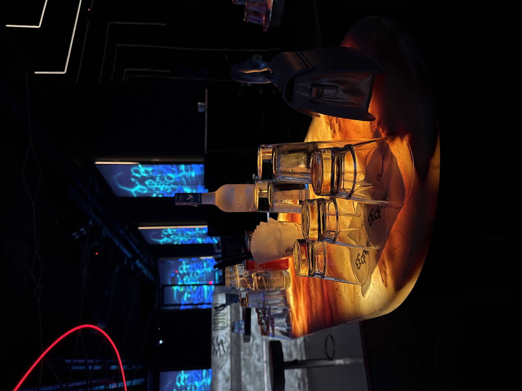 This indoor scene depicts a bar setting, possibly in Sofia, Bulgaria. In the foreground, several stacked glass bottles are on a lit bar surface, positioned to the right. The bottles have a logo printed on them. Behind the bottles is a bottle of alcohol, possibly vodka. Also, napkins and straws.
In the background are illuminated screens displaying blue patterns, and illuminated structures. The lower left of the scene shows a red light in an arc shape. The surrounding environment is dimly lit, with a dark ceiling and the screens as the primary light sources.