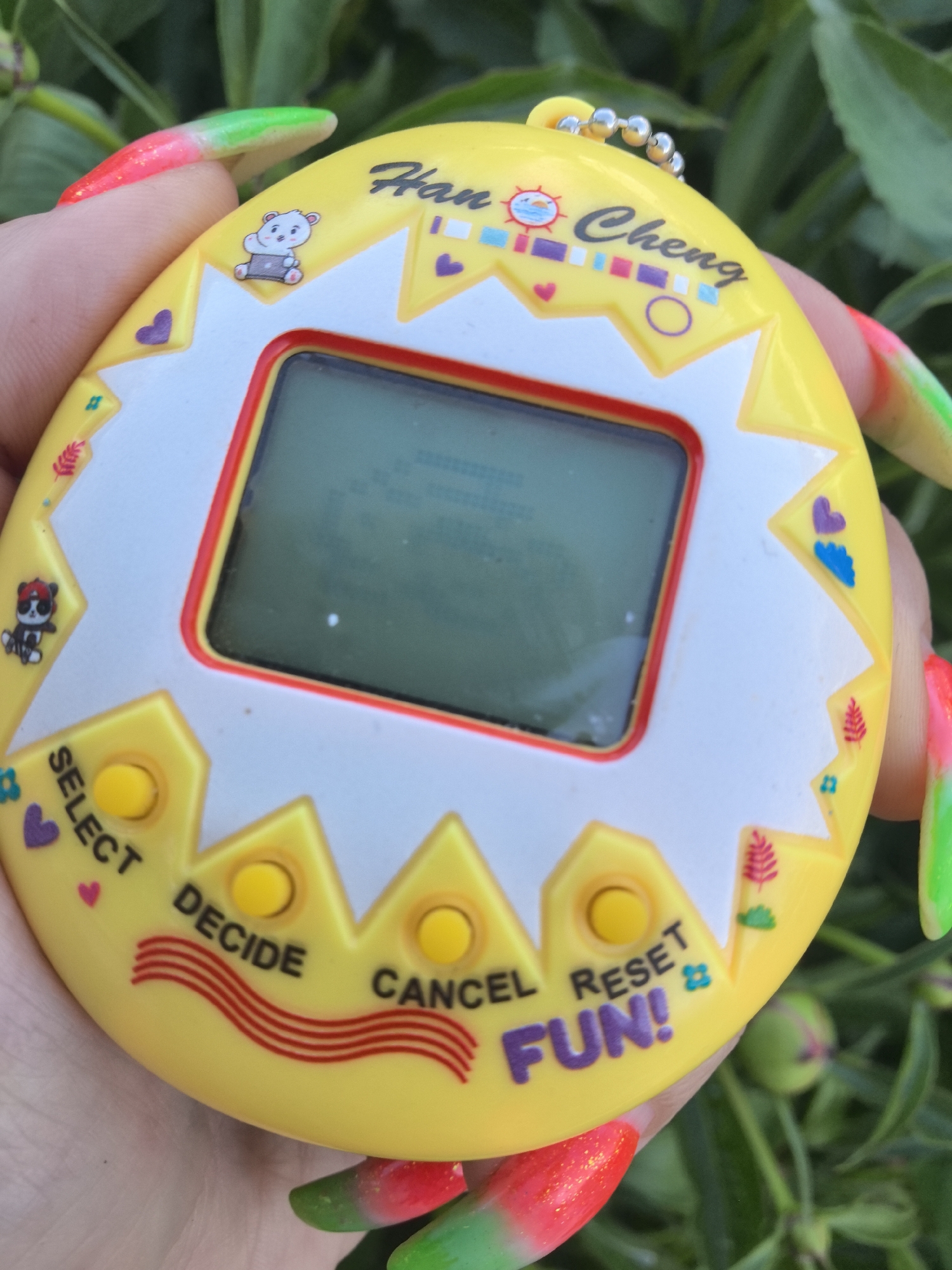 A hand holds a yellow "Han Cheng" virtual pet device with a red-rimmed screen against a background of green foliage. The device has three yellow buttons labeled "SELECT," "DECIDE," "CANCEL," and "RESET." There are various cartoonish decorations on the device, including a bear, hearts, and plants. The nails on the hand are long and painted with a red and green gradient. The location is not specified in the city of Noviy, Russia.