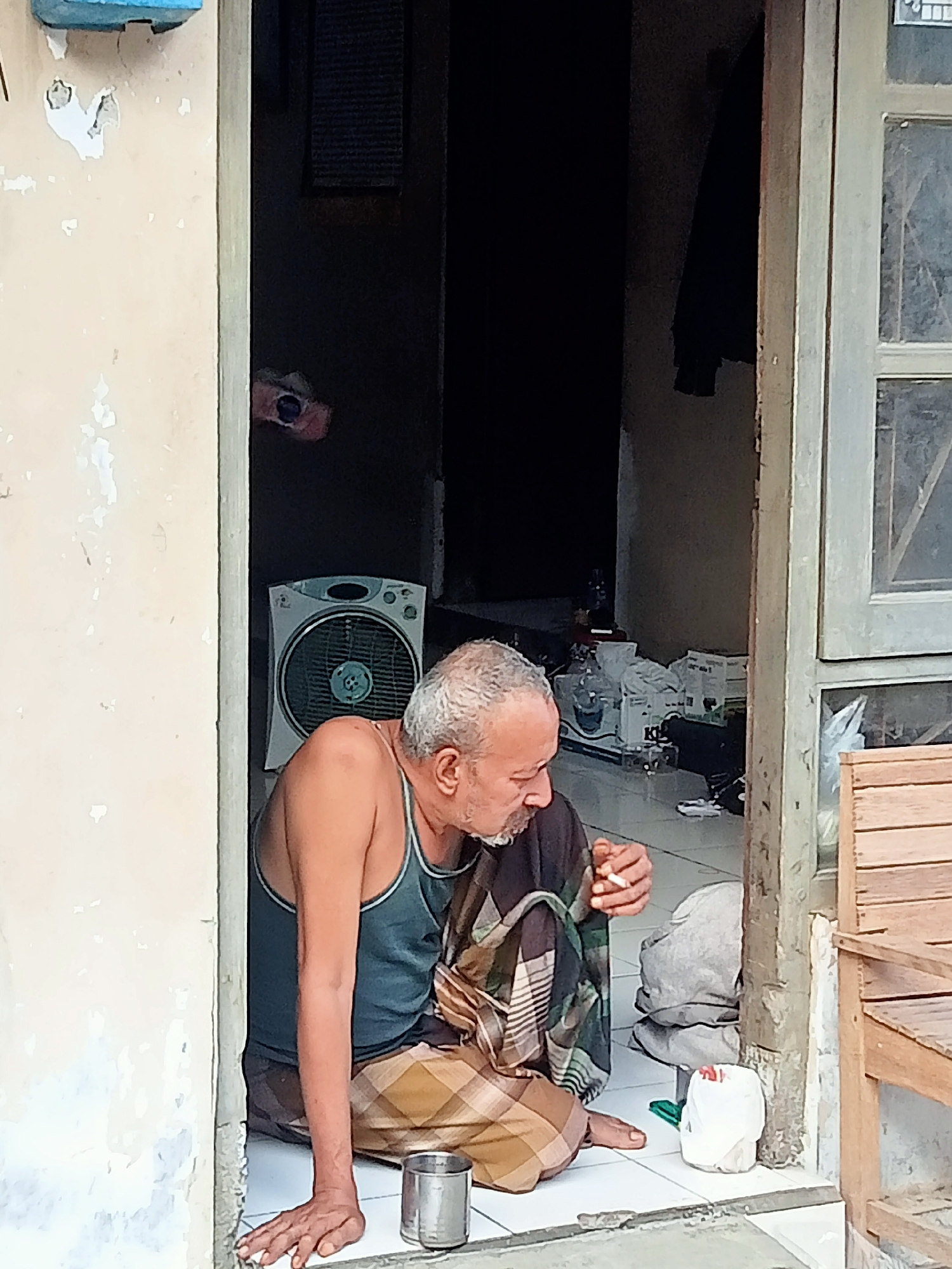 In Kecamatan Pati, Indonesia, an elderly man with graying hair and a beard sits on the floor inside a doorway, smoking a cigarette. He is wearing a dark tank top and a sarong with a checkered pattern. His left hand is raised to his mouth, holding the cigarette, while his right hand rests on the floor. To his right, on the tiled floor, is a metal cup and a white plastic bag. In the background, a white electric fan is visible. The doorway opens to a dimly lit interior with boxes and a black garment hanging on the wall. To the right of the doorway, a wooden chair is partially visible. The walls of the building appear weathered and painted a light beige.