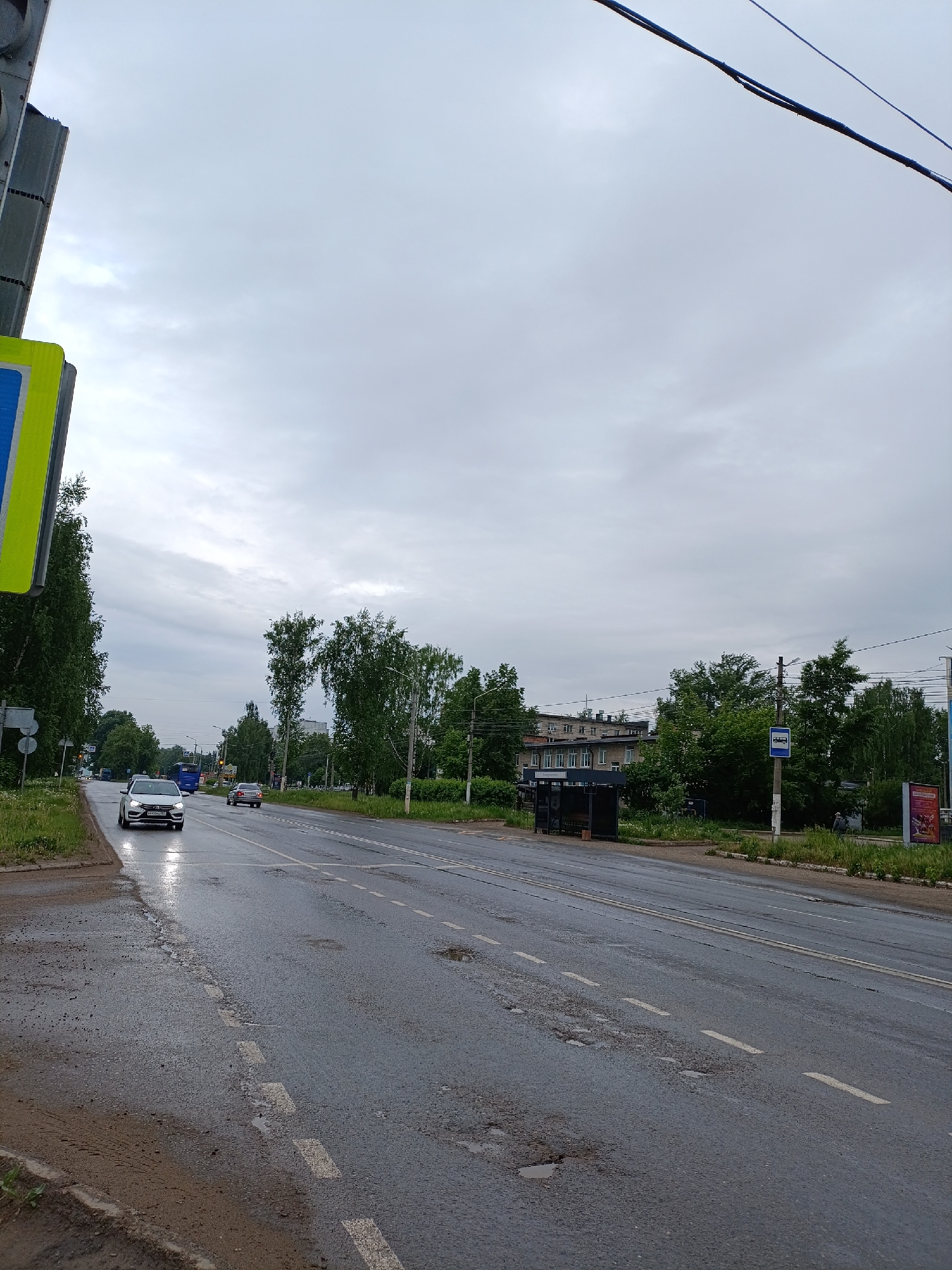The image depicts a street scene in Chaikovsky, Russia on an overcast day. The road is wet, with potholes and dashed lane markings. A white car is visible on the left side of the road, with another car further down the street. Trees line both sides of the road, with buildings visible in the background. A bus stop shelter is present on the right side of the road, along with a bus stop sign. Power lines are visible overhead.