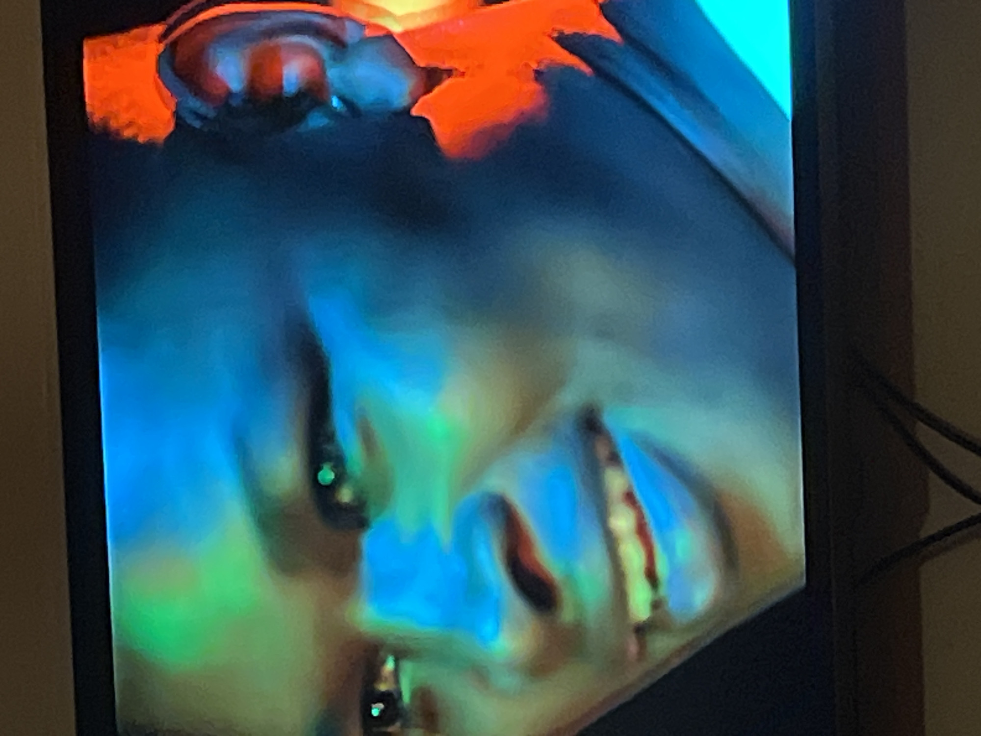 The media file displays a close-up, distorted image of a person's face, likely illuminated by artificial, colorful lighting. The lighting casts vibrant hues of orange, blue, and green across the face, creating abstract patterns and obscuring fine details. The person's eyes are visible, with one appearing to have a green reflection or glint. The overall effect is surreal and somewhat unsettling due to the intense colors and the partial obscuration of the face. The image appears to be captured on a screen, possibly a television or monitor, given the slight blurriness and the presence of a dark frame on the right side. There is no discernible text, weather, or clear indication of the time of day or specific location within the frame itself. The context provided, "Gura Top, Nigeria," suggests the origin or subject matter, but this cannot be visually confirmed from the image alone. The activity or event taking place is not evident; the image is a static portrayal. The emotions conveyed are ambiguous due to the distortion and lighting, but the intense colors could suggest a dramatic or artistic presentation.