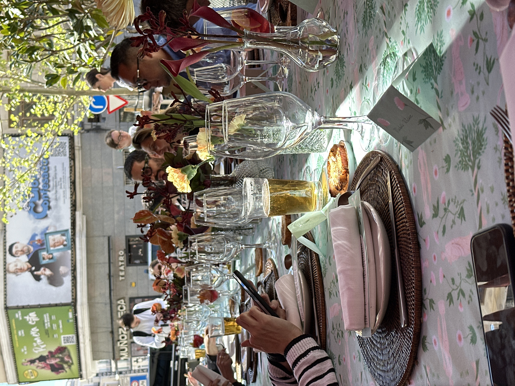 The image captures an outdoor dining scene in Madrid, Spain, with a long table set for a meal. The table is covered with a patterned tablecloth and features elaborate floral arrangements in vases. Multiple wine glasses and beer glasses are arranged along the table. Place settings consist of wicker chargers, plates, napkins, and cutlery. Guests are seated at the table, with some visible in the background and one person in the foreground holding a mobile phone.
In the background, across the street, is the Teatro Muñoz Seca, a theater in Madrid. Advertising posters, including one for "Primera Cita," are displayed on the theater's facade. A street sign and trees are also visible in the background. The scene suggests a special event or celebration taking place in an urban setting.