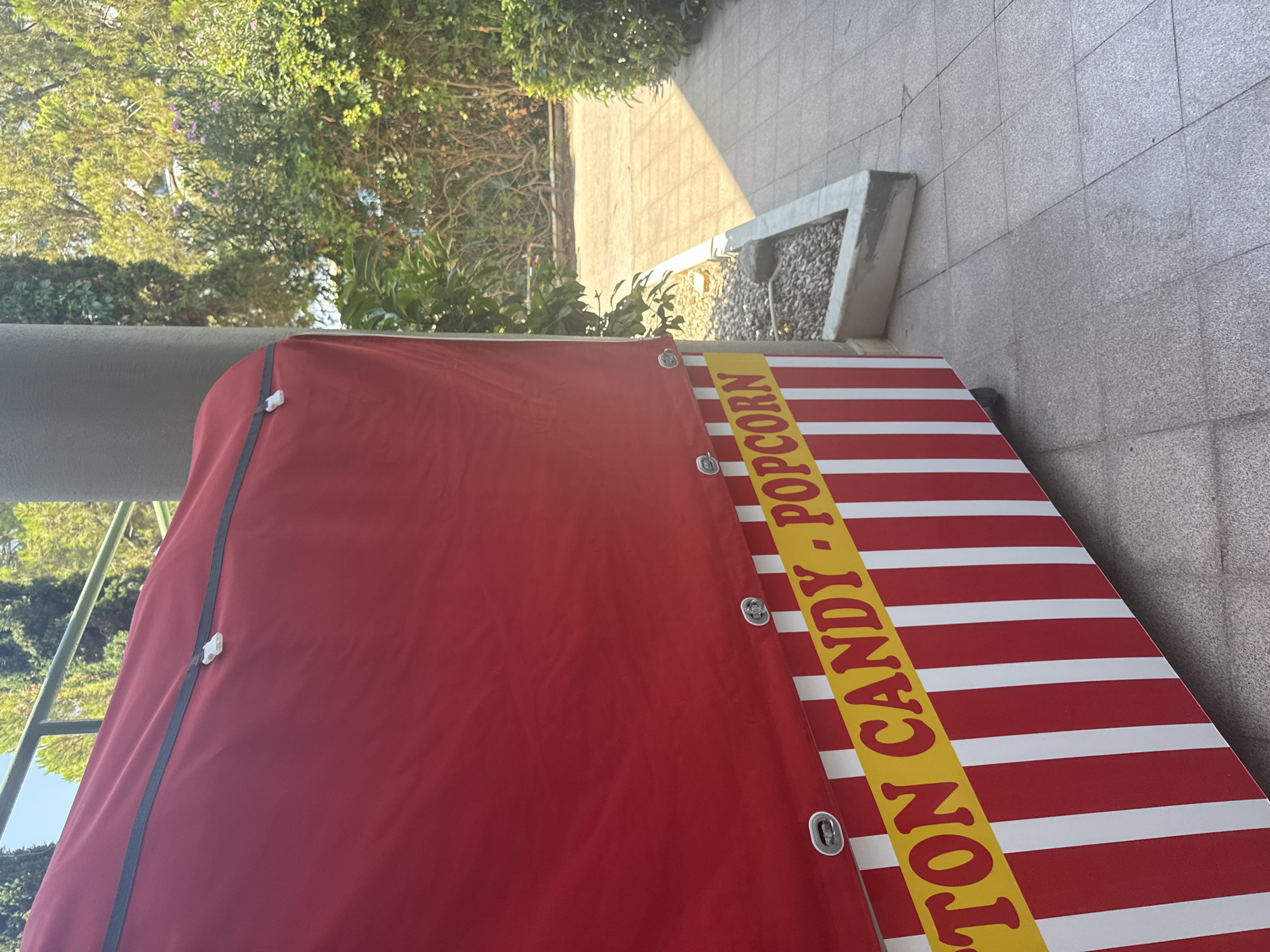 A red carnival-style cart with white stripes is parked on a paved walkway outdoors, with greenery and trees in the background. The cart has "COTTON CANDY POPCORN" written in yellow lettering on a yellow band. It is a sunny day, and shadows are cast on the ground. The cart appears to be closed. There are no people visible in the image. The scene is likely in a public area, possibly a park or promenade, given the paved walkway and manicured landscaping. The cart itself is positioned to the left of the frame, taking up approximately two-thirds of the image.