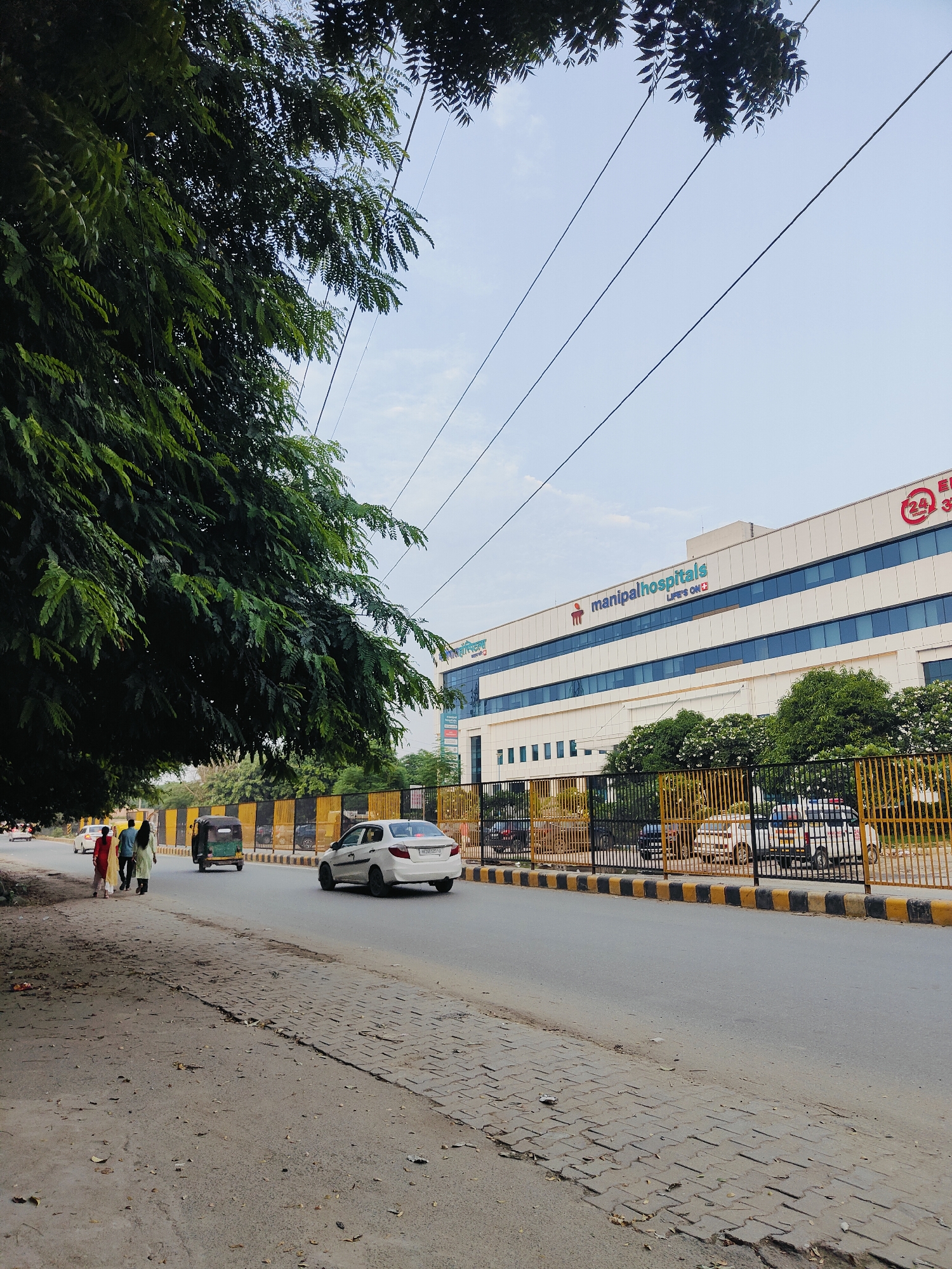 The image shows a street scene in Gurgaon, India, with a large modern hospital building in the background. On the left, a large tree with dense green foliage shades the sidewalk and a portion of the road. The road is paved and appears to be a multi-lane thoroughfare. Several vehicles are visible on the road, including a white sedan in the foreground, an auto-rickshaw behind it, and other cars further down the street. A yellow and black striped barrier lines the side of the road in front of the hospital. The hospital building is a multi-story structure with a white facade and numerous blue-tinted windows. The "manipalhospitals" logo is prominently displayed on the building. A few people are walking on the sidewalk on the left side of the road. The sky is overcast with scattered clouds. The overall scene depicts a typical urban environment in India.