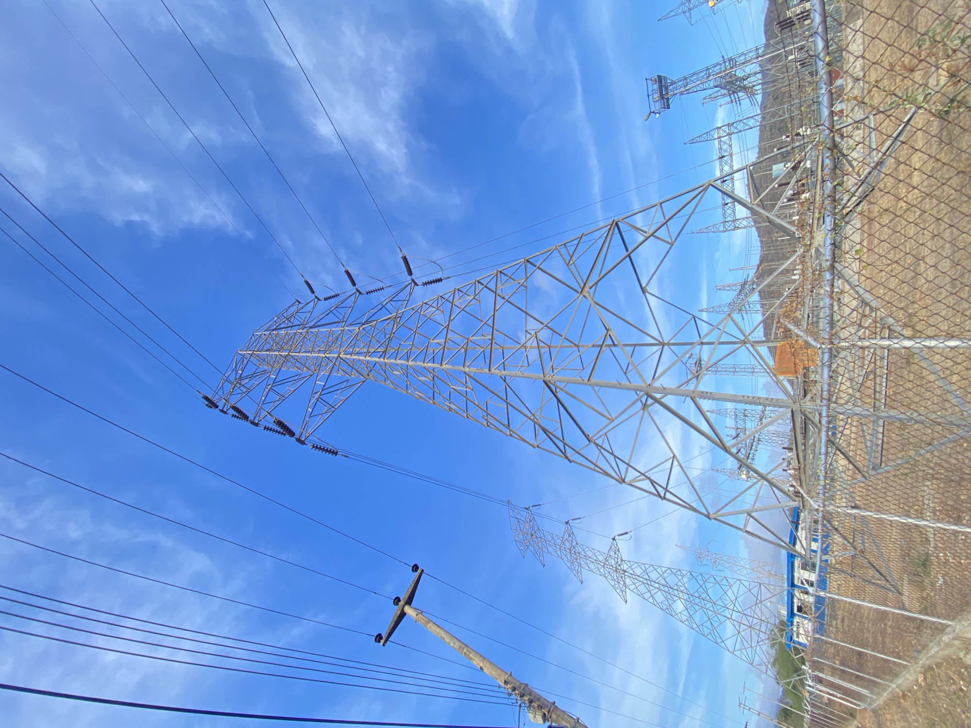 This image captures an extensive electrical power transmission and substation facility under a bright blue sky with scattered wispy white clouds. The scene is dominated by multiple high-voltage electricity transmission towers, primarily of the lattice steel type, stretching across the landscape.
In the foreground and central to the image, a large, tall transmission tower supports numerous power lines, clearly visible with their insulator strings. To the lower left, a concrete utility pole with cross-arms and several attached wires further indicates the presence of electrical distribution infrastructure.
The right side of the image reveals an electrical substation enclosed by a chain-link fence. Within the fenced area, various pieces of electrical equipment such as transformers, insulators, circuit breakers, and busbars are discernible. A blue and white building is also visible behind the substation equipment. Beyond the substation and the fence, a range of dry, undulating hills or mountains extends into the background, hinting at a natural, possibly rugged, landscape.
The time of day is clearly daytime, characterized by bright, clear lighting. The weather is fair with good visibility. The prominent electrical infrastructure, combined with the natural background of hills, suggests an essential utility installation in a rural or semi-rural setting. No legible text is visible on any of the structures or equipment within the frame.