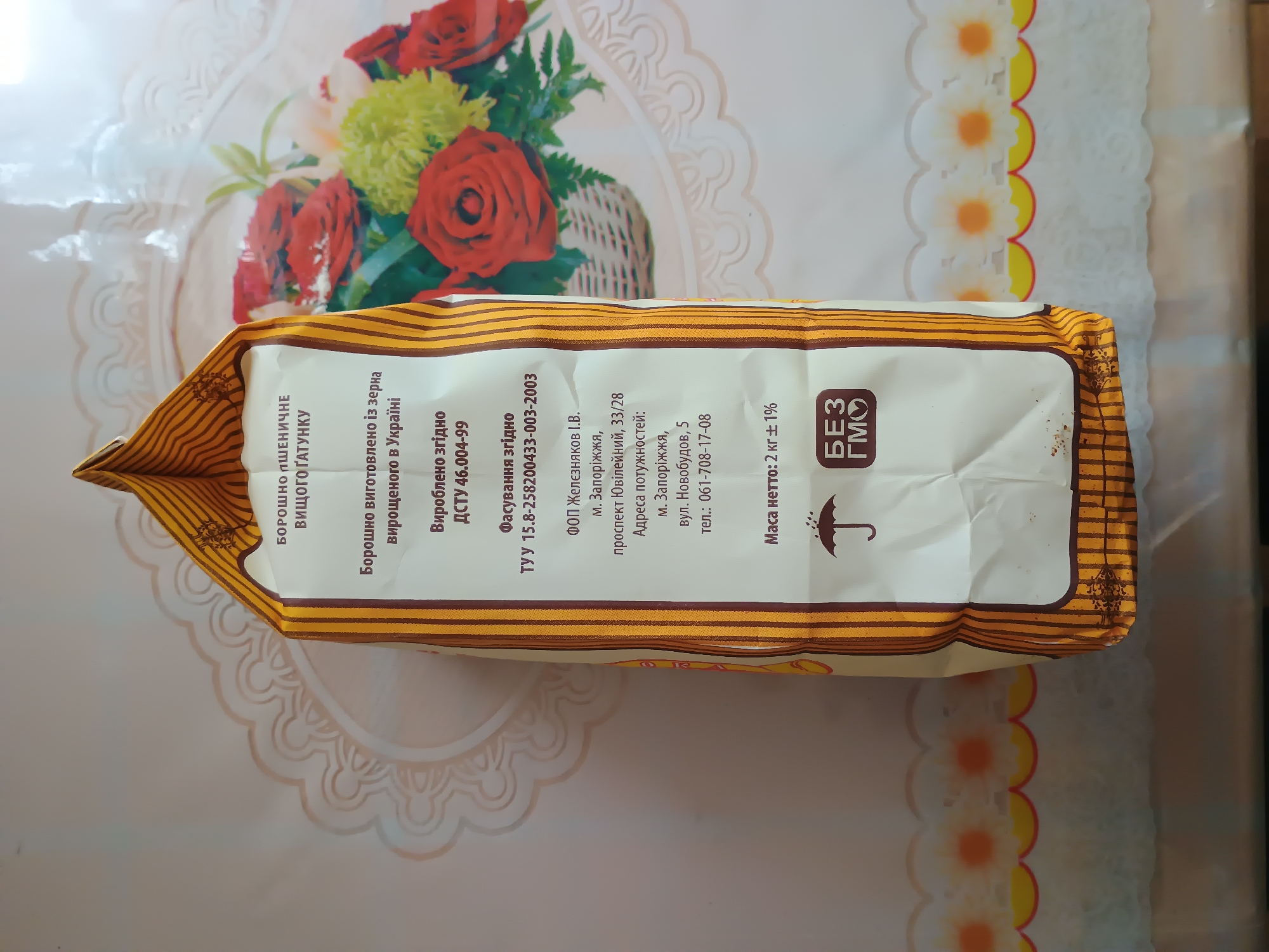 The image shows a close-up of a bag of wheat flour, likely a product from Ukraine. The bag is oriented horizontally and is placed on a tablecloth with a floral and scalloped edge pattern. The dominant colors of the packaging are white and yellow, with brown stripes and text.
Visible text on the bag includes:
- "БОРОШНО ПШЕНИЧНЕ ВИЩОГО ГАТУНКУ" (Wheat flour of the highest grade)
- "Борошно виготовлено із зерна Вирощеного в Україні" (Flour made from grain grown in Ukraine)
- "Вироблено згідно ДСТУ 46.004-99" (Produced according to DSTU 46.004-99)
- "Фасування згідно ТУУ 15.8-258200433-003-2003" (Packaging according to TUU 15.8-258200433-003-2003)
- Manufacturer details: "ФОП Желєзняков І.В.", "м. Запоріжжя, проспект Ювілейний, 33/28", "Адреса потужностей: м. Запоріжжя, вул. Новобудов, 5", "тел.: 061-708-17-08" (Indicating the manufacturer is a private entrepreneur, located in Zaporizhzhia, Ukraine, with specific street addresses and a phone number).
- "Маса нетто: 2 кг ± 1%" (Net weight: 2 kg ± 1%)
- A "БЕЗ ГМО" (GMO-free) logo with an umbrella icon.
The background is a tablecloth with a decorative pattern featuring flowers and scalloped edges, suggesting a domestic or retail setting. The lighting appears to be natural or soft indoor lighting. The image has a slight tilt. There are no people visible in the image. The focus is entirely on the product and its packaging information.