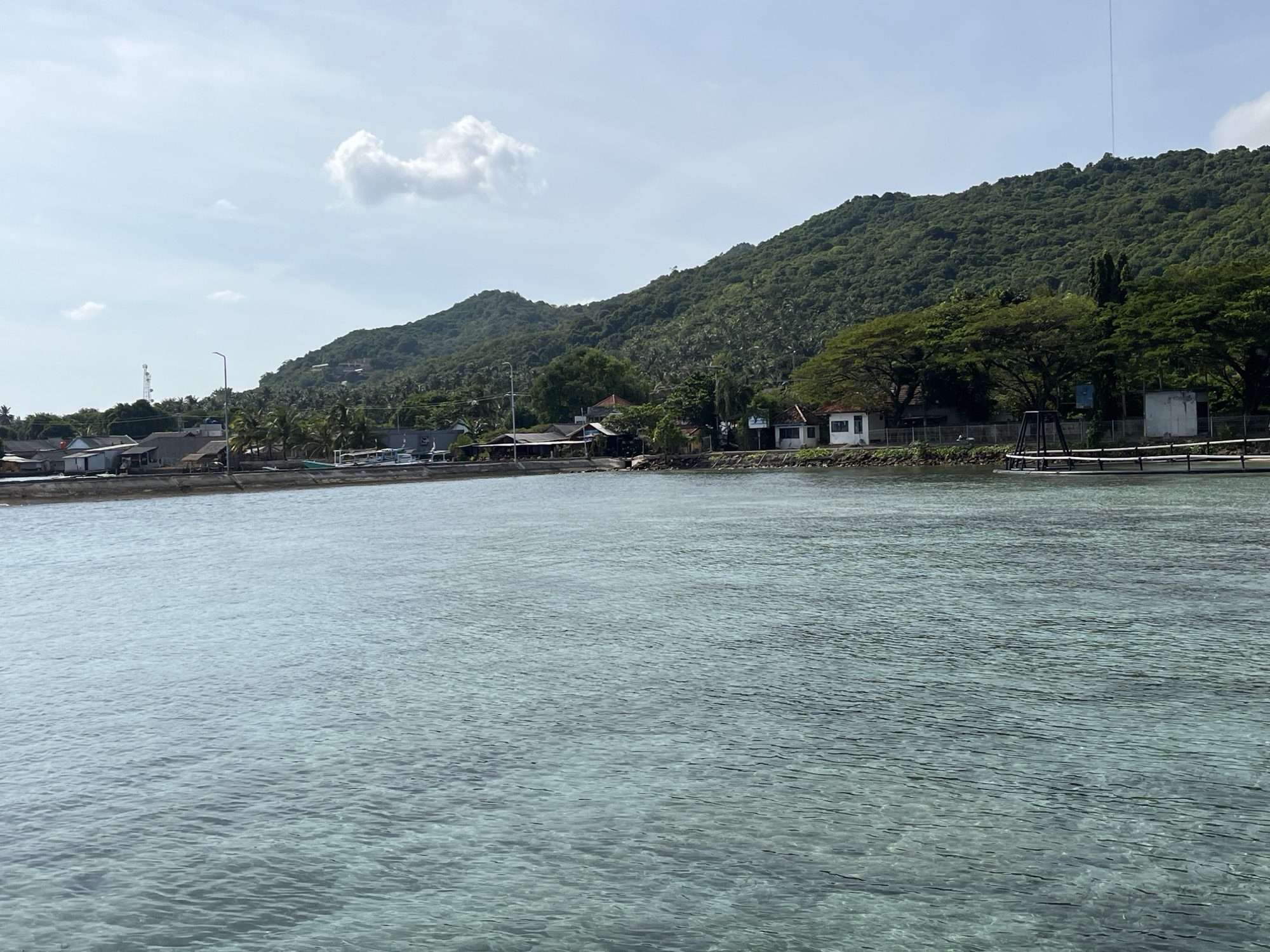The image depicts a serene coastal scene in Karimunjawa, Indonesia. The foreground is dominated by crystal-clear turquoise water, revealing the sandy seabed below. The water is calm, with gentle ripples reflecting the sunlight. Along the shore, a small village is visible, characterized by a cluster of houses with red-tiled roofs and some buildings with white facades. Several boats are docked near the shore. Behind the village, a lush green, densely forested hill rises steeply, suggesting a tropical island environment. The sky is a light blue with a few scattered white clouds. A tall telecommunications tower can be seen on the far left, and a long pier or boardwalk extends into the water on the right side of the image. The overall impression is one of a peaceful, tropical coastal settlement.