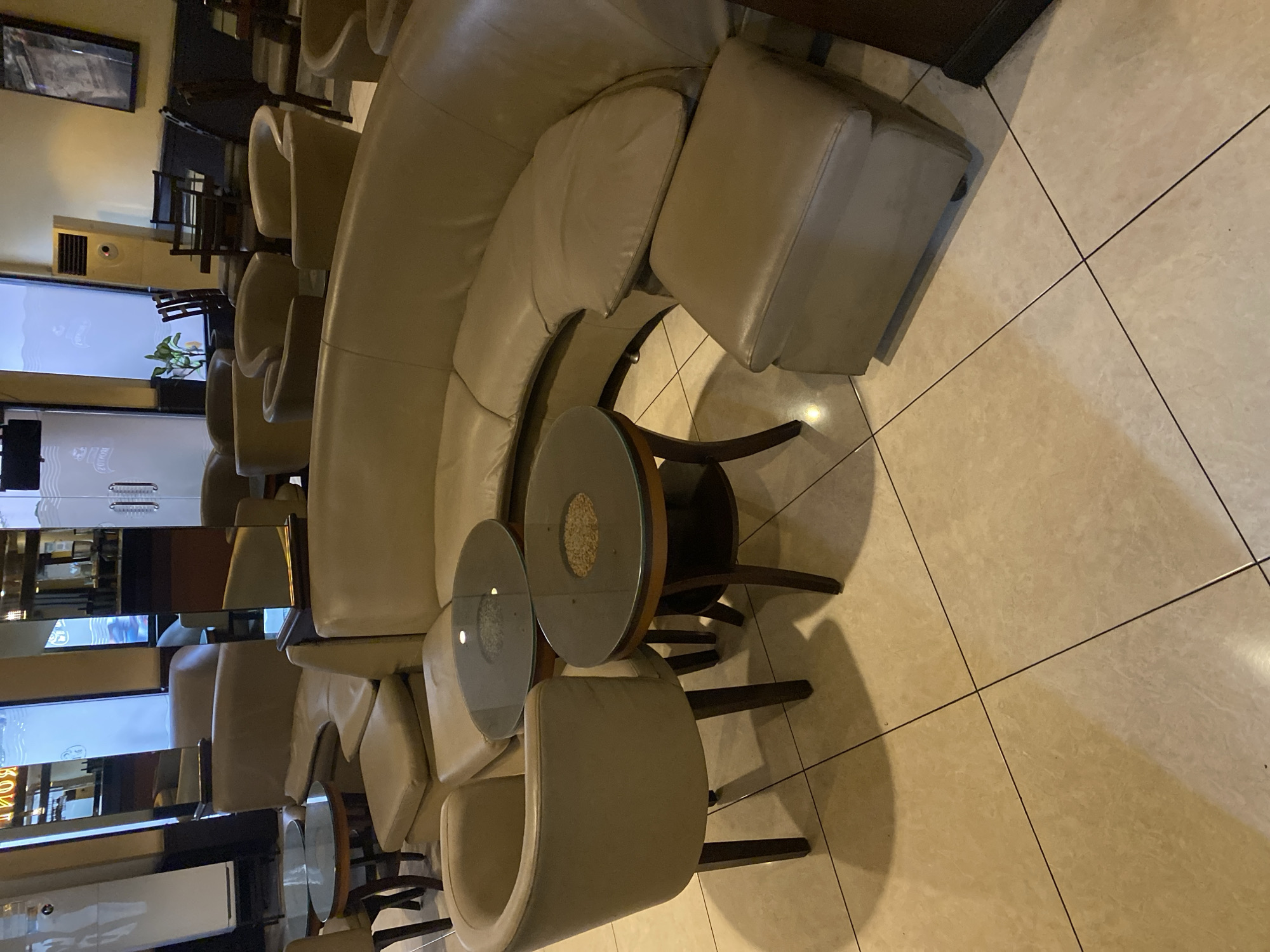 The image depicts a seating area in what appears to be a cafe or lounge. The main subject is a large, curved, light brown leather sofa with an ottoman. In front of the sofa are two round tables with dark glass tops and wooden bases. To the left of the sofa, a single light brown leather chair with a curved back is positioned near one of the tables. The flooring is made of light-colored square tiles. In the background, reflections in mirrors show more seating areas, tables, and what looks like a window with text. An air conditioning unit is visible on the wall in the background. The overall impression is that of an indoor commercial space. The image does not contain any specific visual cues to indicate it is located in Kediri City, Indonesia.