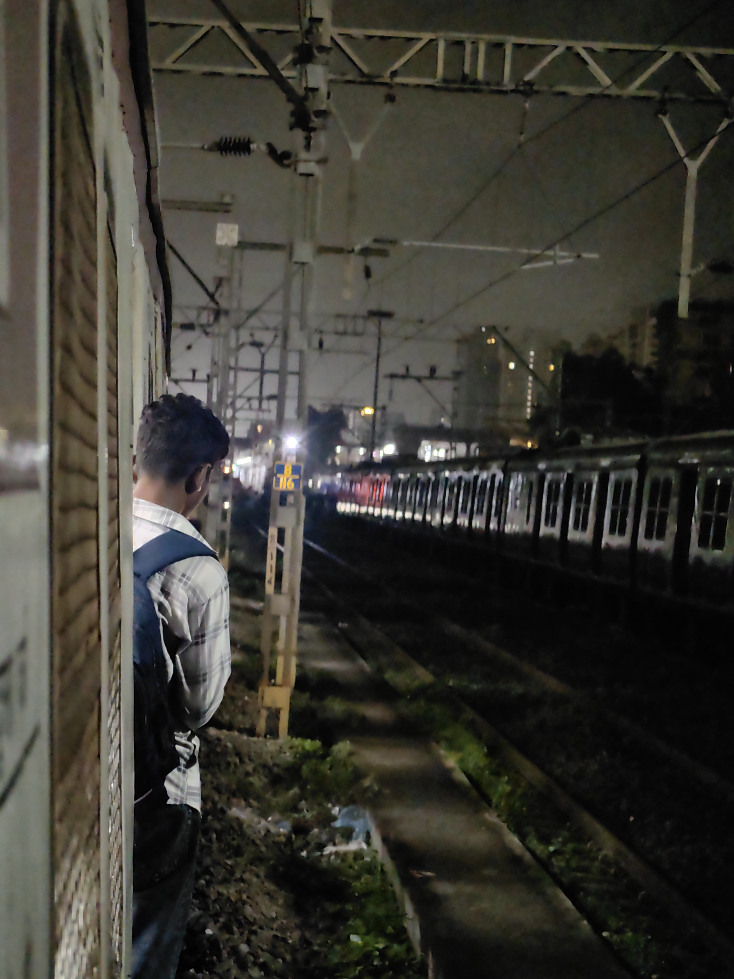 A young male wearing a plaid shirt and a backpack stands on a train platform at night, looking away from the camera towards a train on the adjacent tracks. The train appears to be at a standstill. The platform is illuminated by artificial lights, and buildings are visible in the background, suggesting an urban environment. The scene is likely a railway station in Mumbai, India, given the presence of local trains and the characteristic overhead electrical infrastructure commonly seen in the city's suburban railway network. The ground on the platform is covered with gravel and some vegetation.