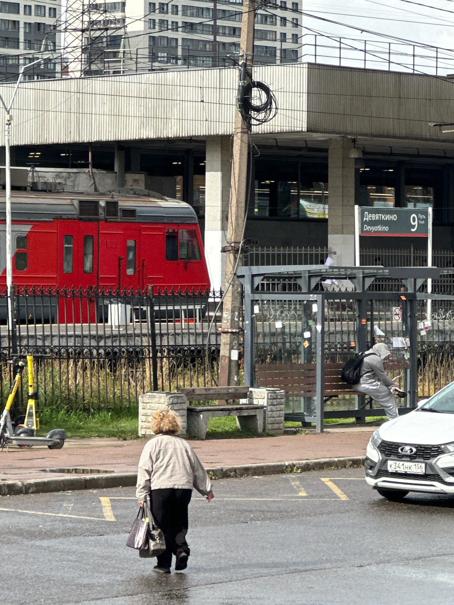 An elderly woman with curly blonde hair, wearing a light-colored jacket and black pants, walks away from the camera on a wet asphalt road. She carries two bags in her left hand. In the background, a red train is stopped at a station with a sign that reads "Devyatkino 9". A person in a gray hoodie and sweatpants sits on a bench at a bus stop, looking down. A white car is parked on the right side of the road. A yellow electric scooter is visible on the left side of the image. The scene appears to be in a city, possibly Vsevolozhsk, Russia, given the signage.