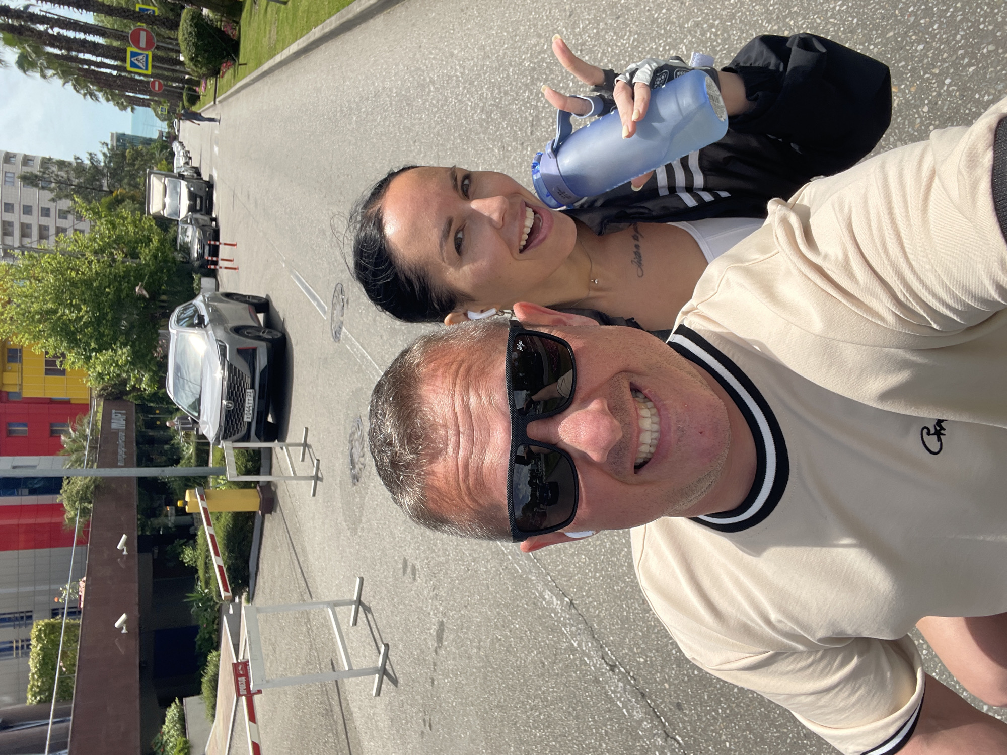 A medium eye-level, color outdoor selfie shows a man and woman. The man, in the foreground, has short gray hair, tan skin, sunglasses, and a beige t-shirt with black and white neck and sleeve trim. The woman, behind him, has black hair, tan skin, is smiling, and holding a blue water bottle. She is wearing wireless earphones, a white tank top, and a black jacket with white stripes on the sleeves. The background shows a street and buildings, with parked cars, trees, and traffic signs. The street is asphalt. There are white and red barriers present. The building on the left has the word "MART" written in Cyrillic characters. The location is the city of Sochi, Russia.
