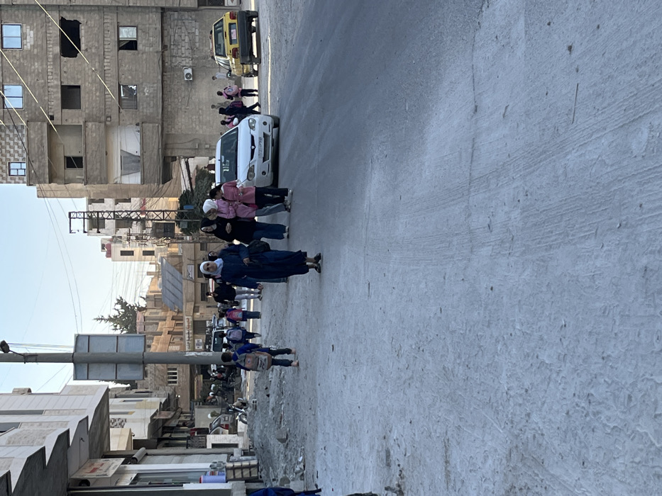 This image depicts a daytime street scene in Mu'adammiyat ash-Sham, Syria.
In the foreground, the ground on the left consists of uneven, dusty terrain with scattered debris and small piles of dirt. This transitions into a paved asphalt or concrete road surface on the right, exhibiting minor cracks and dust.
Approximately 15-20 individuals are present. In the midground, slightly left of center, a female adult wearing a dark blue abaya and a light-colored headscarf walks towards the camera, holding a dark bag. To her right, a group of three female adults walk together; one wears black, and two wear pink tops. Further down the street, multiple children, most carrying backpacks, are visible walking on both sides of the road, suggesting they are school-aged. A young boy with a light-colored backpack is walking away from the camera on the unpaved left side. Other adults are present in the background.
Vehicles include a white sedan positioned on the right side of the road, behind the group of women, and a yellow taxi cab parked further down the street on the right.
Multi-story buildings constructed from concrete blocks and masonry line both sides of the street. Structures on the left exhibit signs of partial construction or disrepair, with exposed rebar and unfinished sections. Buildings on the right display worn facades, darkened window openings, and one external air conditioning unit. A prominent utility pole with attached signage stands on the left side of the paved road. The sky above is clear.