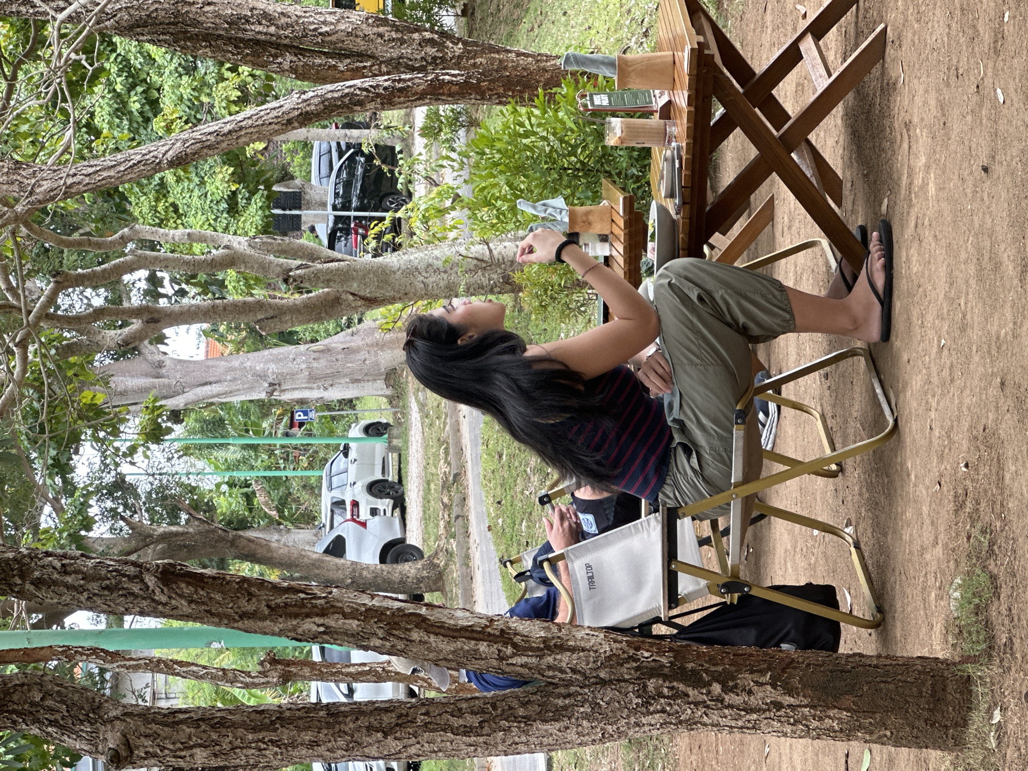 In a shaded outdoor seating area, likely at a cafe in Kuta Selatan, Indonesia, a woman with long dark hair sits at a wooden table, facing away from the camera and looking to her right. She is wearing a striped top and olive green pants, with black flip-flops on her feet. She holds a fork in her right hand, poised near a plate of food. On the table, a glass of iced coffee, a menu, and a small wooden holder with napkins are visible.
Behind her, to the left, another person is seated in a folding camping chair. They are wearing a blue shirt and a baseball cap, and their hands rest on the chair's armrests. A black backpack is hung on the back of their chair.
The seating area is surrounded by trees with rough bark, and in the background, several cars are parked, including white SUVs and a black sedan. The ground is covered in dirt and some scattered leaves. The overall scene suggests a casual dining experience in an outdoor environment.