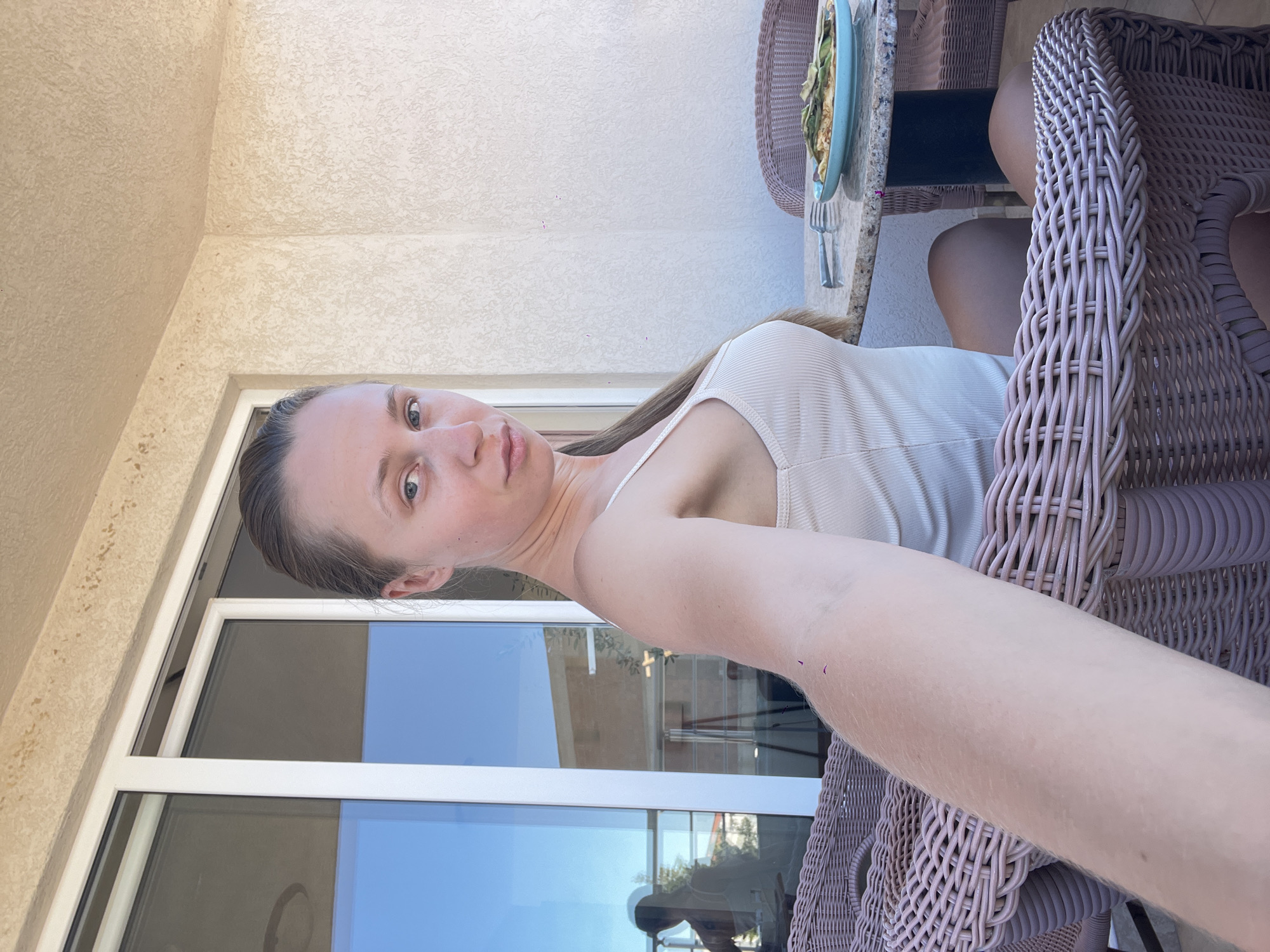 A young woman with blonde hair pulled back is seated on a wicker chair on a patio. She is wearing a light-colored tank top and looking at the camera. In front of her, on a granite-topped table, is a blue plate with food and a fork. Behind her is a large glass sliding door that reflects the sky and buildings. The woman's arm is extended towards the camera, as if taking a selfie. The environment appears to be a balcony or patio area, likely in a warm climate, with a textured beige wall and ceiling overhead.