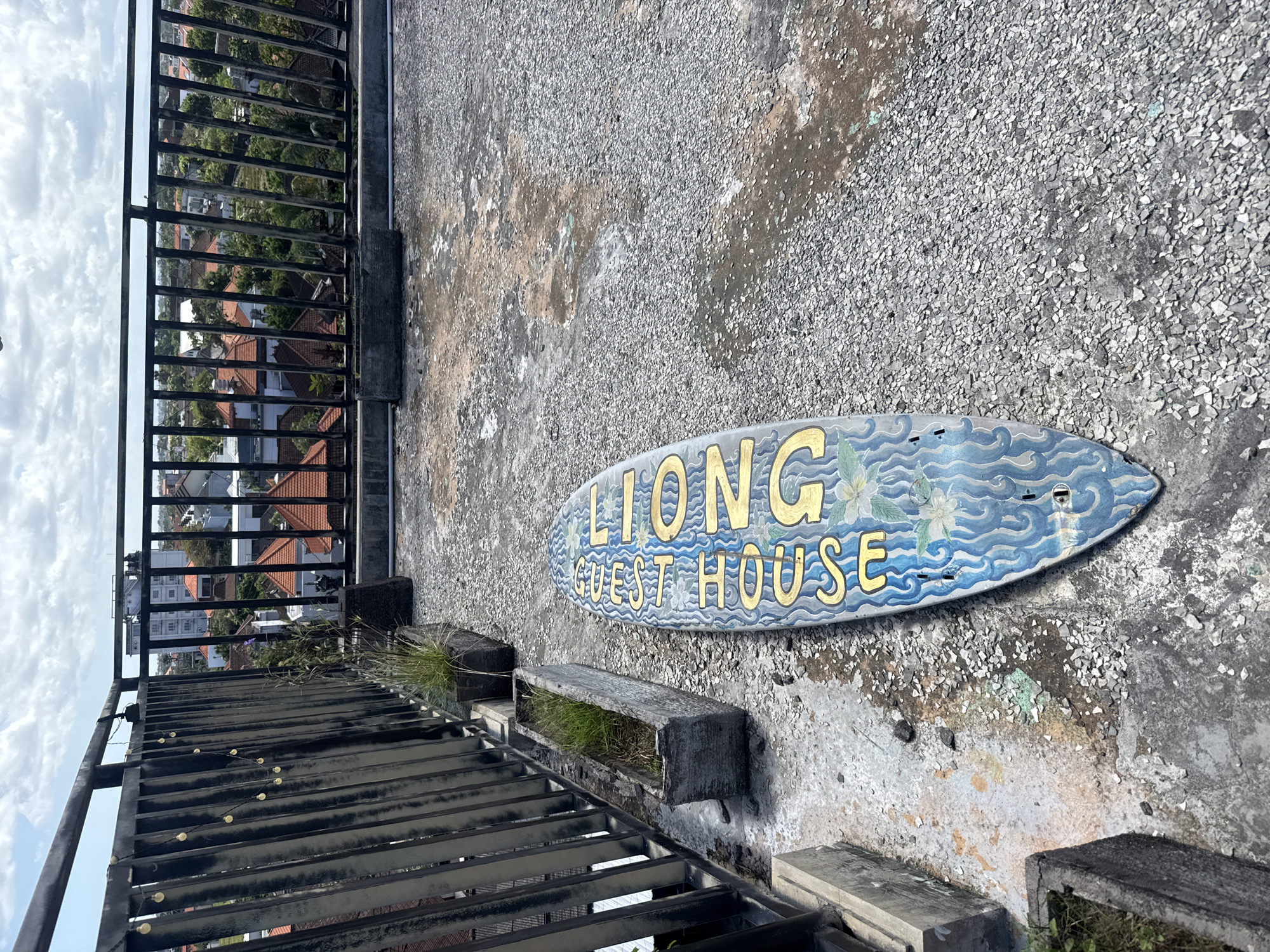 On a gravel rooftop in North Kuta, Indonesia, lies a weathered sign reading "LIONG GUEST HOUSE" in faded gold letters on a blue, surfboard-shaped background. The sign is slightly chipped and damaged, showing signs of age and exposure. A black metal railing lines the edge of the rooftop, offering a view of the city below, characterized by buildings with red-tiled roofs and lush greenery. Concrete planters with grass are arranged along the railing, showing signs of neglect. The rooftop surface is gray and uneven, with patches of discoloration and wear.
