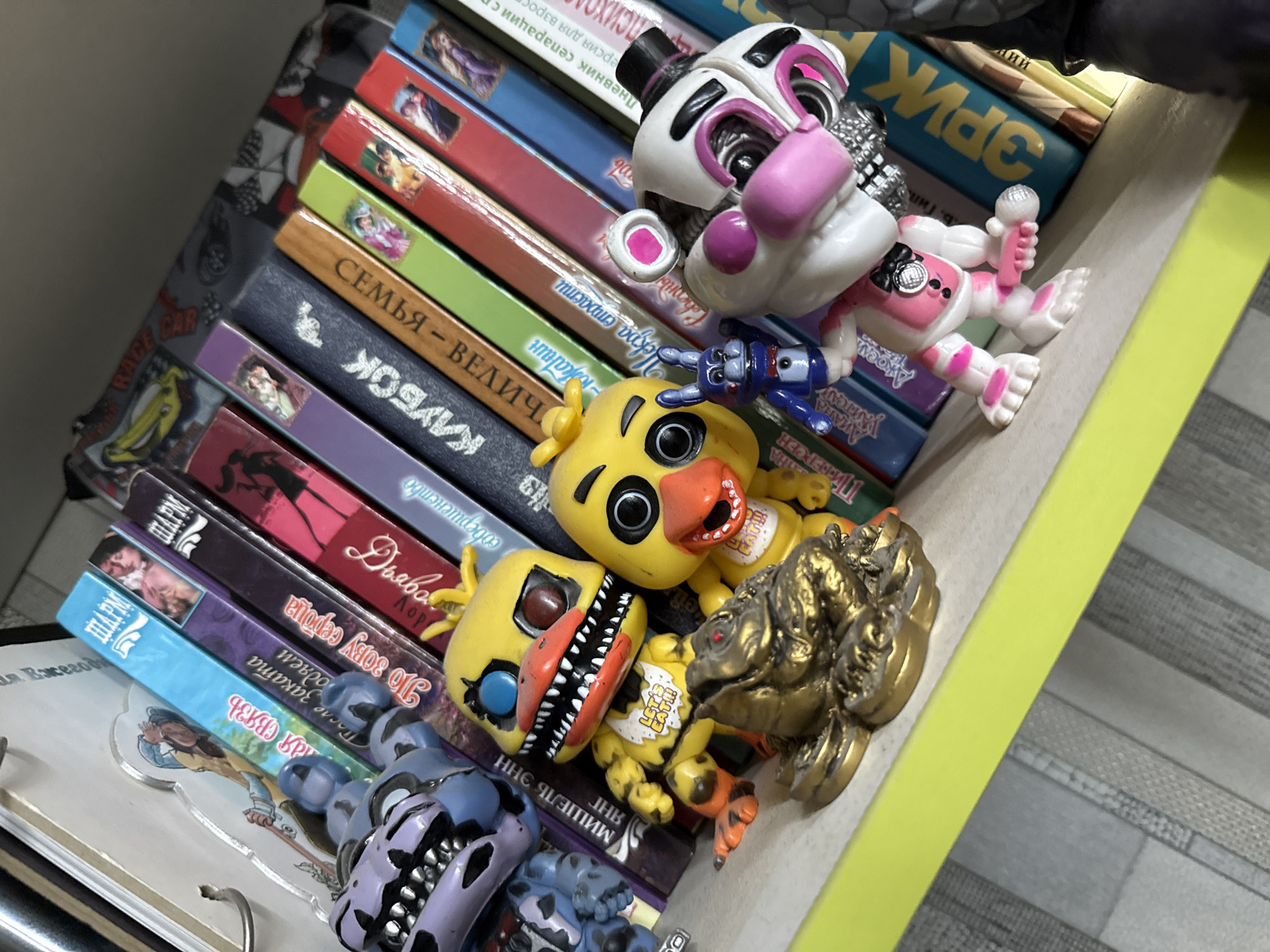 The image shows a shelf with several books and figurines. The books are stacked horizontally and vertically, with titles visible in Cyrillic script. The figurines are colorful and appear to be characters from a popular game or franchise, with one resembling a yellow chicken with sharp teeth, another a white animatronic with pink accents, and a third a small blue rabbit. A gold-colored figurine of what looks like a frog or toad is also present. The shelf itself is a light green color and is positioned against a backdrop of what appears to be a tiled wall. The overall lighting suggests an indoor setting. There is no discernible information in the image that would indicate a specific location in the city of Simferopol, Ukraine.