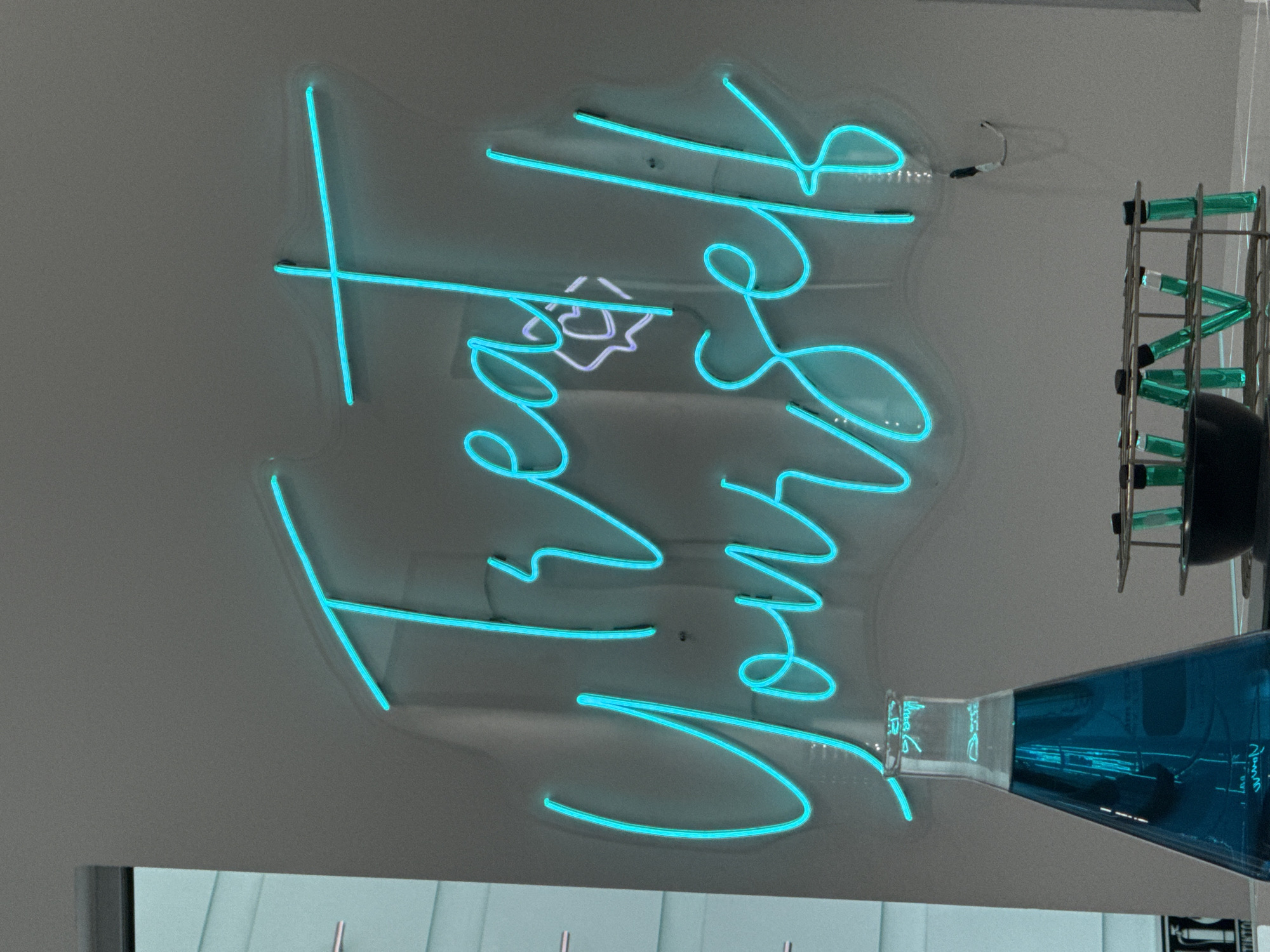 The image shows a neon sign that reads "Trust Yourself". The sign is in a bright cyan color. To the right of the sign is a metal structure with green tubes and a black sphere. A blue, cone-shaped object with writing on it is attached to the bottom of the sign. The background is a plain, light-colored wall. The location is San Bernardino Tlaxcalancingo, Mexico.