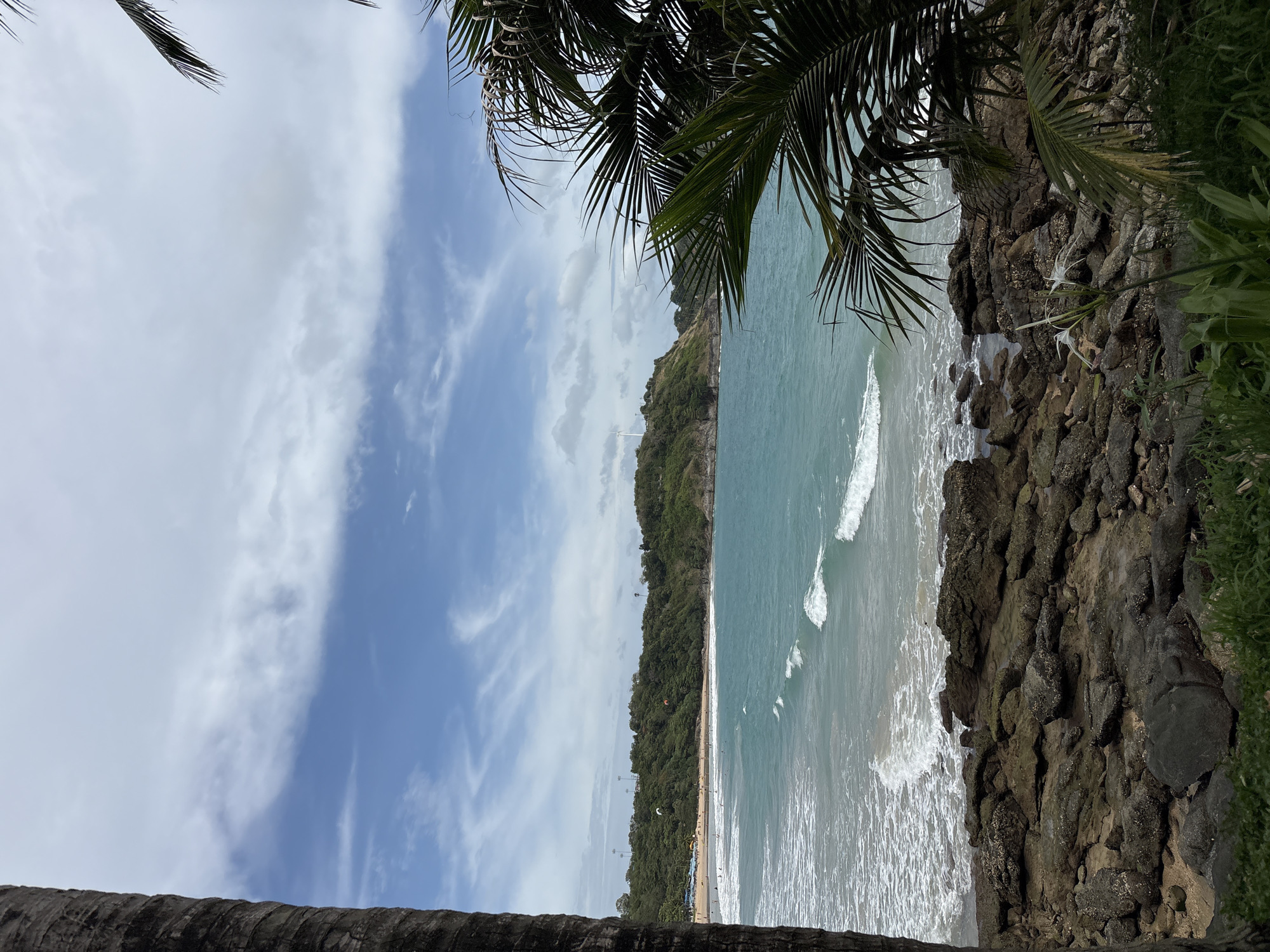 The image presents a view of a beach in Rawai, Thailand. The frame is partially obscured by palm fronds in the upper corners and a palm trunk at the bottom. A rocky outcrop dominates the right side of the image, descending towards the sea. The turquoise ocean meets a sandy beach bordered by a dense forest canopy. The beach is populated with small figures, suggesting people enjoying the coastal environment. Above the beach, the sky is patterned with white and blue clouds.