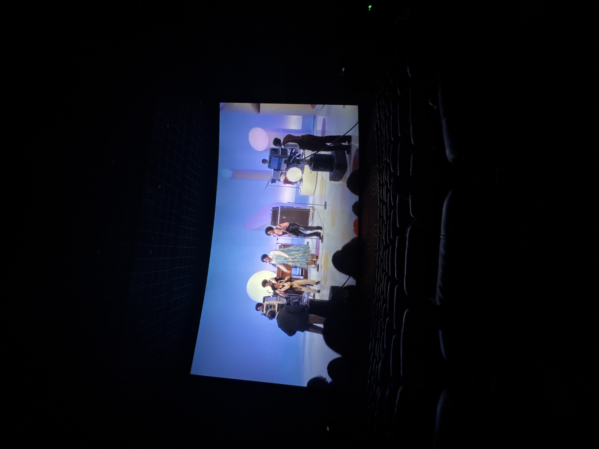 The image shows a movie theater in Taihe District, China. The screen displays a band performing. There are several rows of empty seats in front of the screen. The room is dark except for the light from the screen.