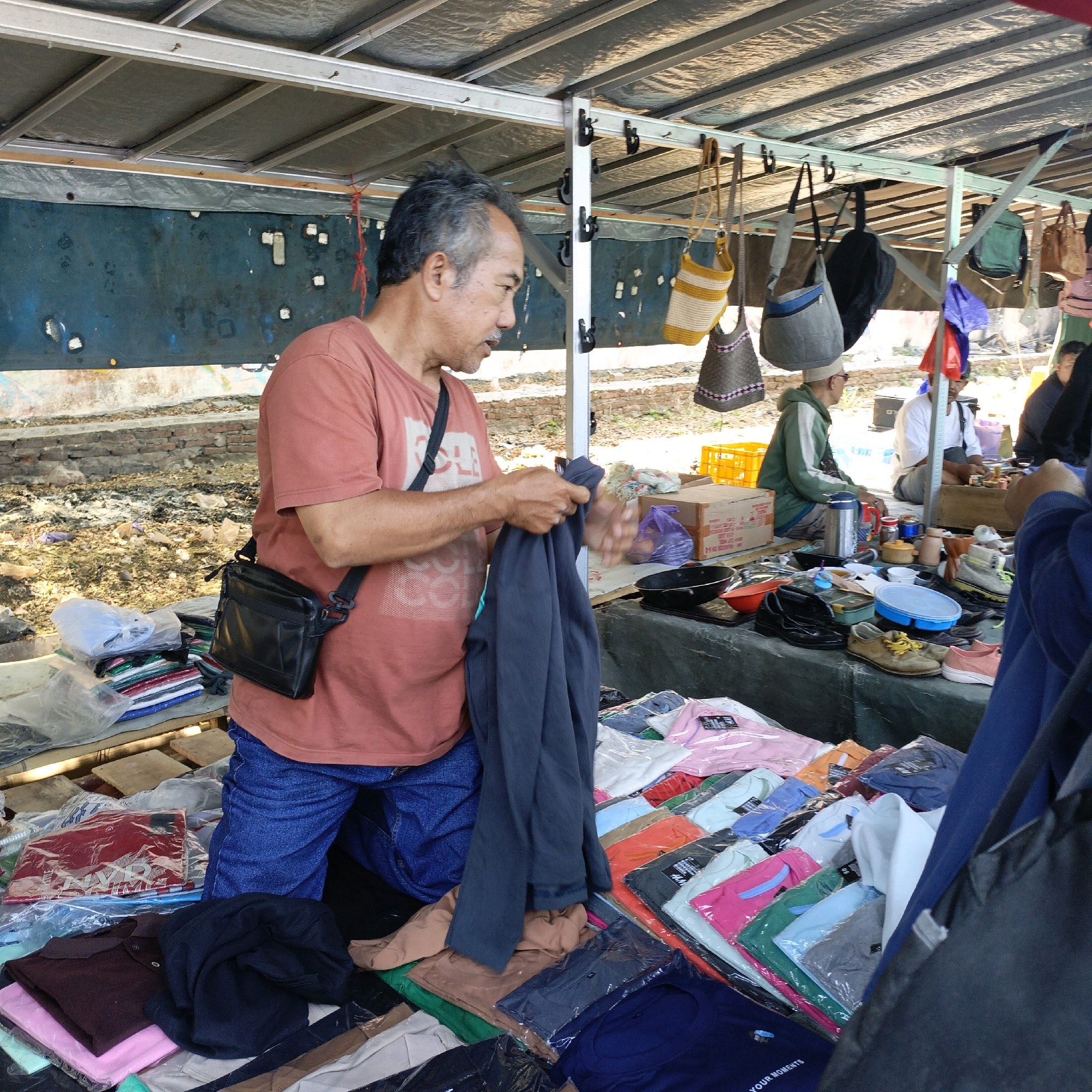 A medium shot captures a market stall scene in Ambarawa, Indonesia. An adult male, with graying hair and a mustache, wearing a salmon-colored t-shirt and blue jeans, stands on the left, holding a dark grey garment. He has a black crossbody bag. In front of him, a table is laden with folded clothes, some in plastic packaging. To his right, a collection of handbags are hung from the stall's roof. Further to the right, other individuals are visible, including one man in a green hoodie and another in a white shirt, sitting behind a display of various items like shoes, bottles, and cooking utensils. The stall is covered by a corrugated metal roof, and the background shows a brick wall and debris.