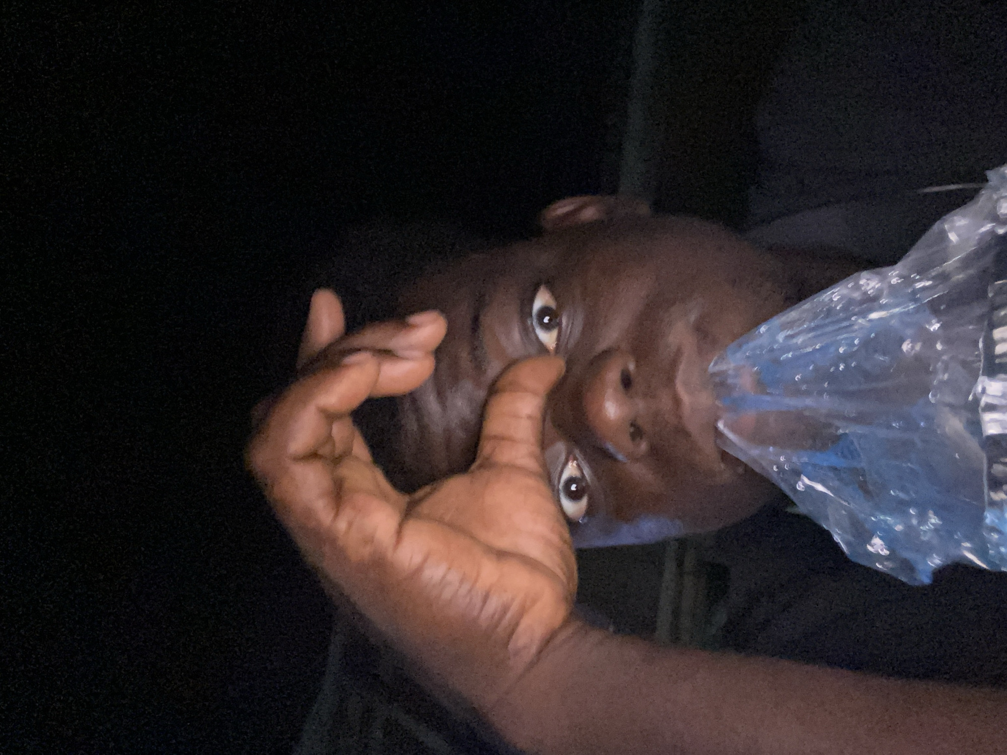 An African-American male is pictured at close range, his face predominantly in focus. He appears to be holding a clear plastic bag to his mouth. One of his hands is positioned near his face, with fingers extended. The background is dark, obscuring environmental details. The location is Unknown, Unknown.