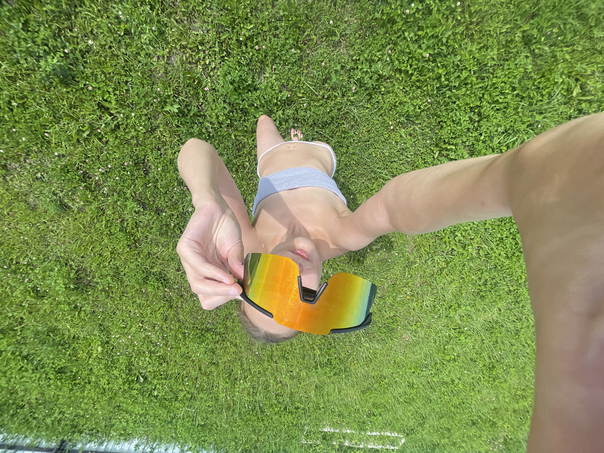A person lies on a grassy lawn, holding a pair of mirrored sunglasses in front of their face. The sunglasses have orange and green reflective lenses. The person is wearing a grey sports bra and white underwear. Their body is angled towards the right of the frame, with their legs extended. The lawn is green and appears to be a park or backyard in the city of Podshivalovo, Russia, with small pink wildflowers scattered throughout. The image is taken from a high-angle, looking down at the person.